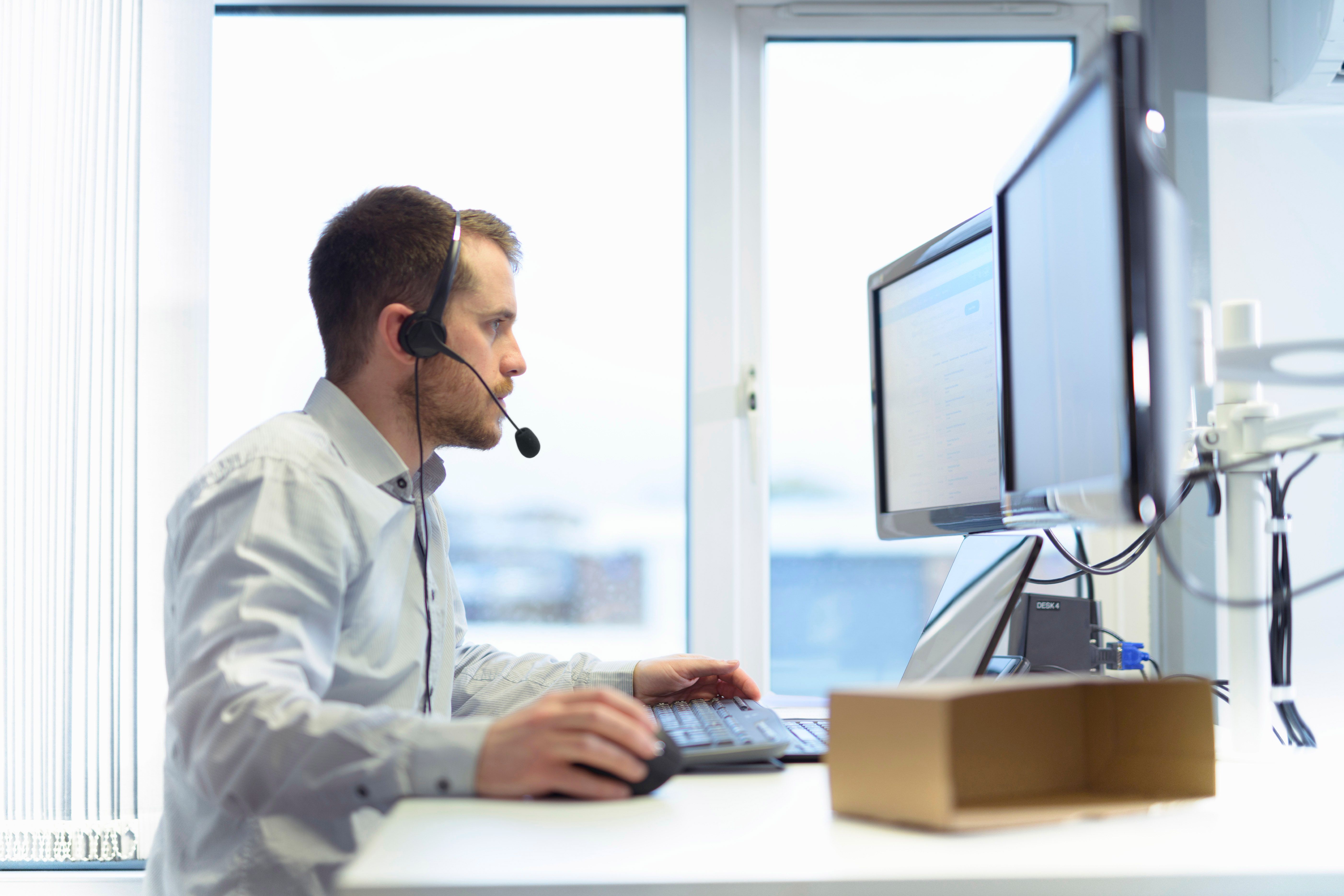A man wearing a headset works at a desk with two computer monitors.