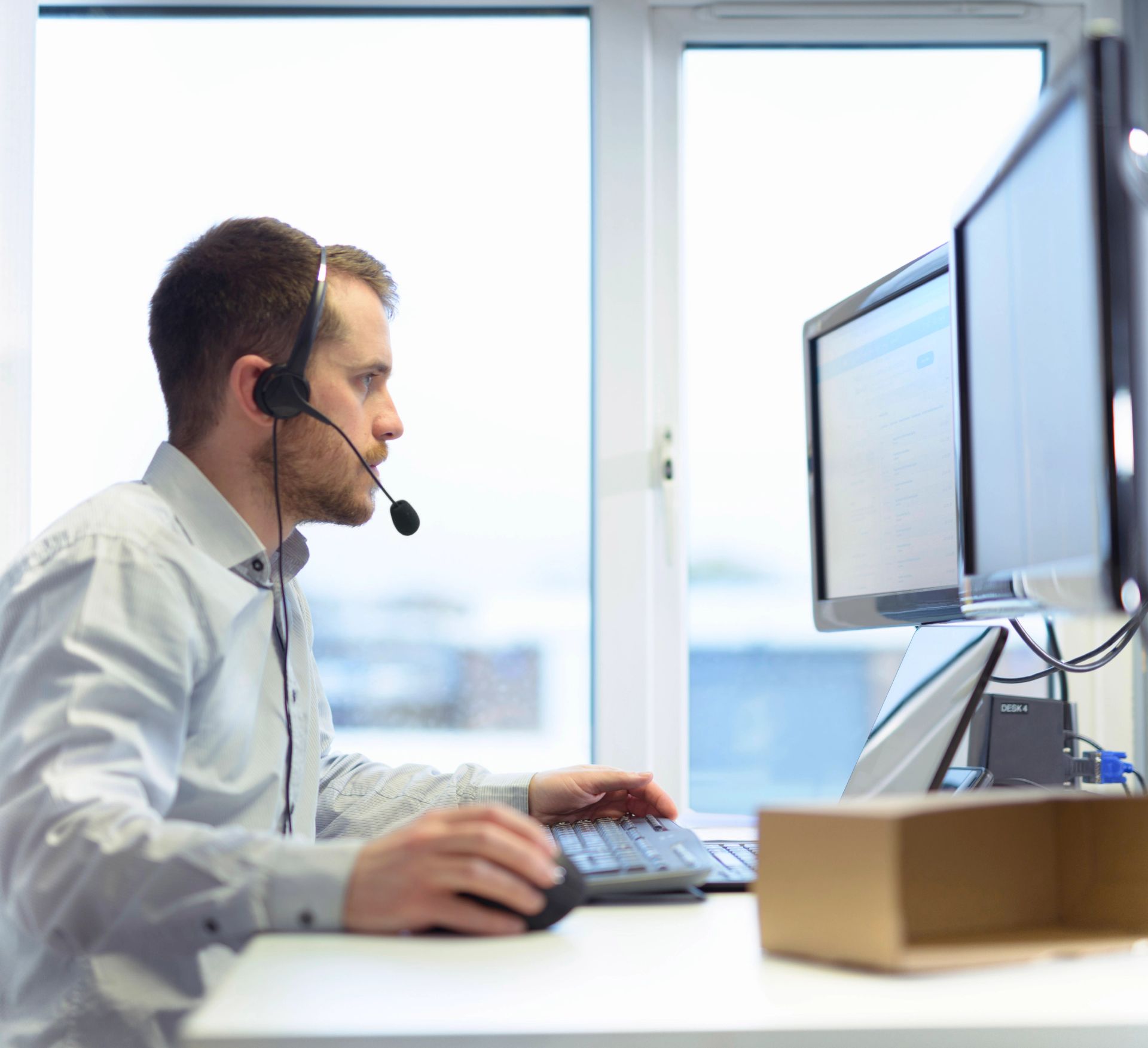 A man wearing a headset works at a desk with two computer monitors.