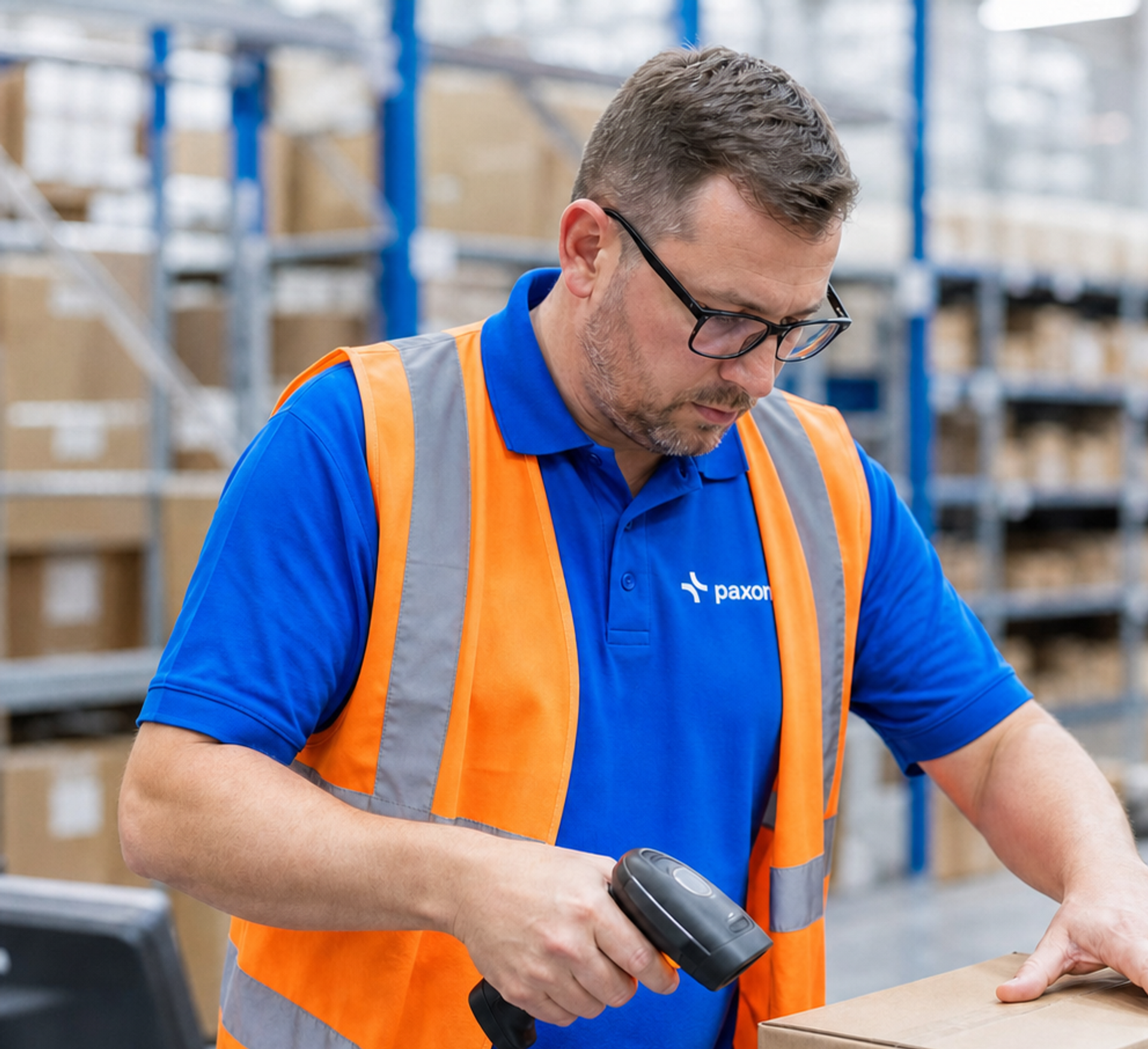 Man in a Paxon shirt and orange safety vest scanning a package in a warehouse.