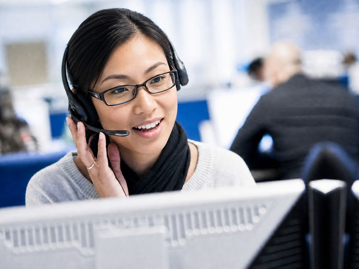 Une jeune femme asiatique souriante, avec casque-micro et lunettes, travaille sur un ordinateur dans un bureau.