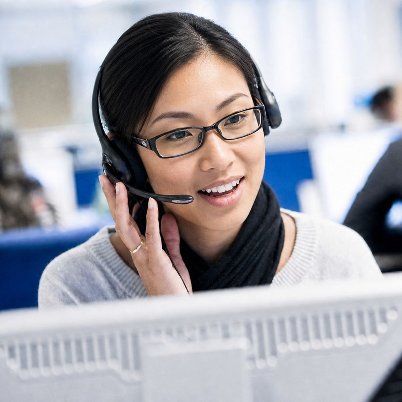 Une jeune femme asiatique souriante, avec casque-micro et lunettes, travaille sur un ordinateur dans un bureau.
