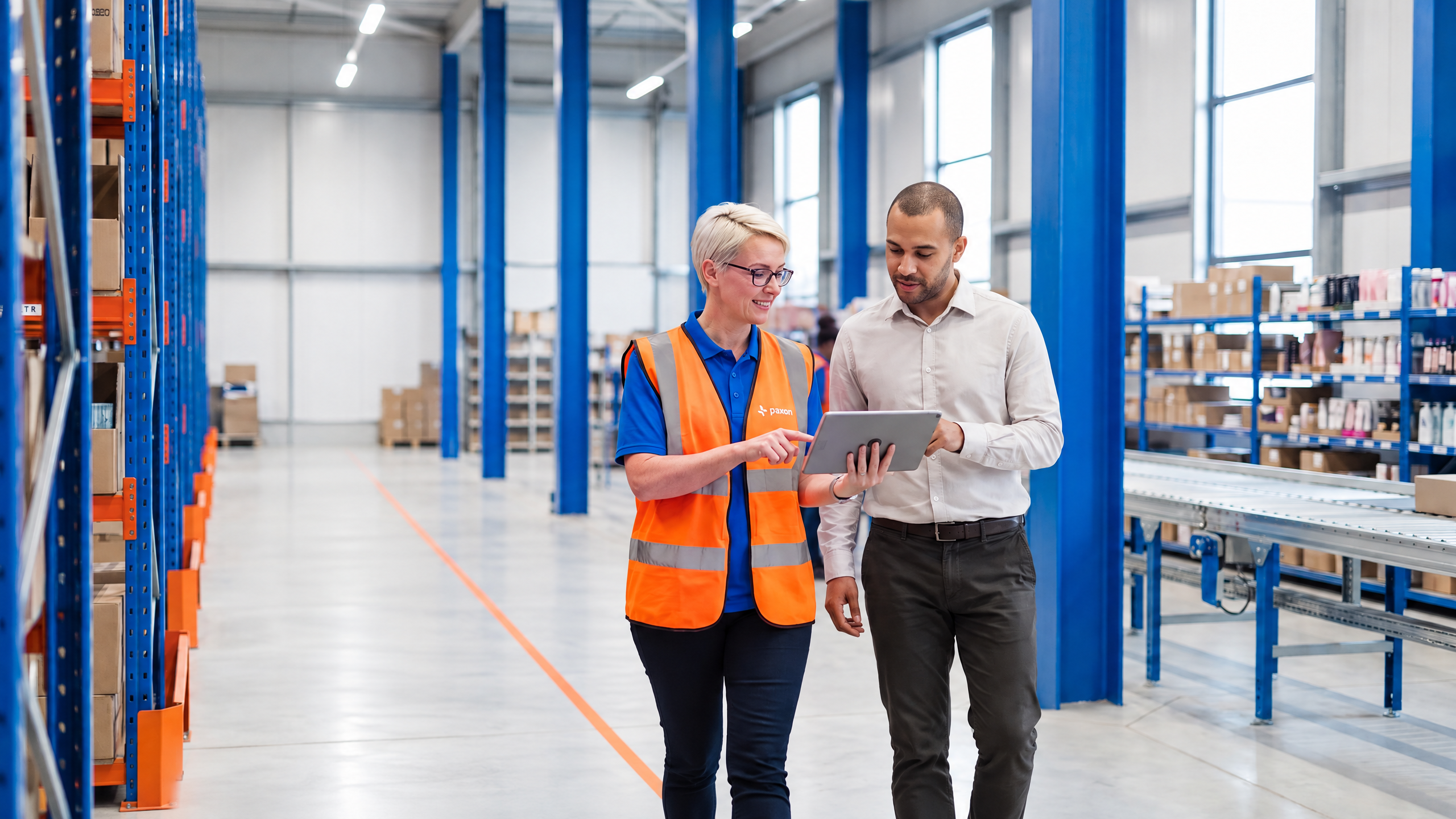 A woman in a safety vest points at a tablet held by a man, in a warehouse.