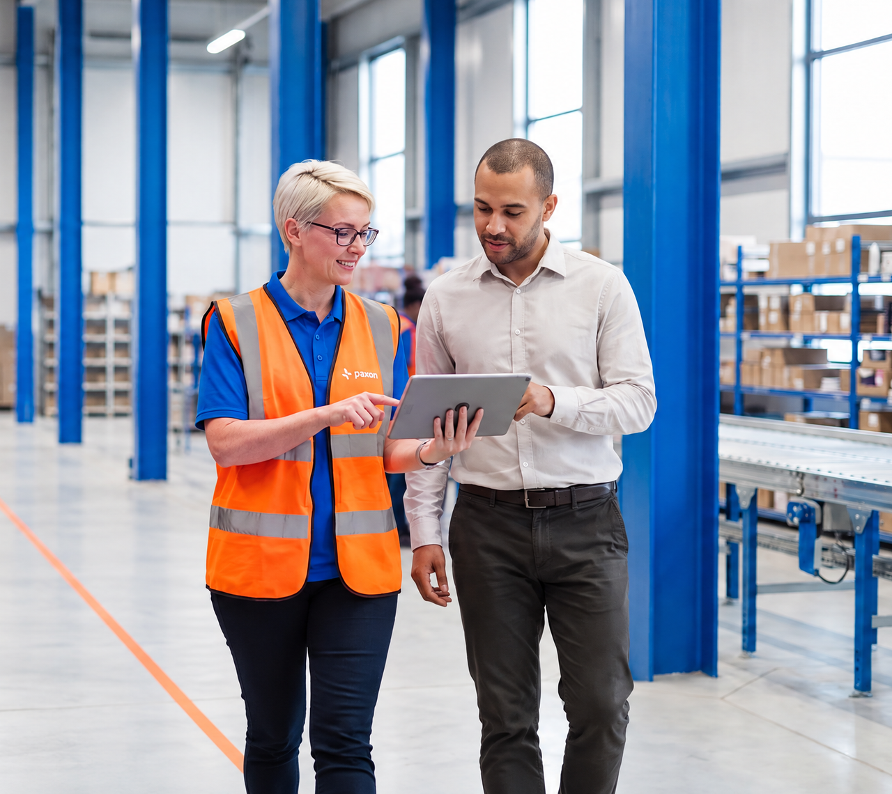 A woman in a safety vest points at a tablet held by a man, in a warehouse.