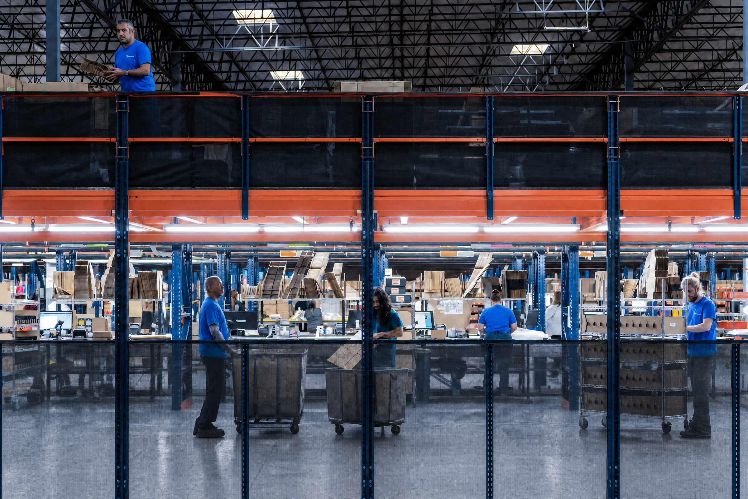 A brightly lit warehouse with workers in blue shirts processing items at tables, surrounded by tall shelves of boxes.