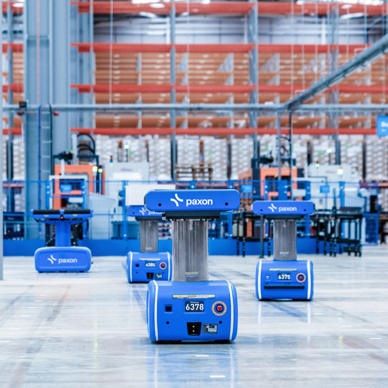 Meerdere blauwe autonome mobiele robots van Paxon op de vloer van een magazijn, met rekken op de achtergrond.