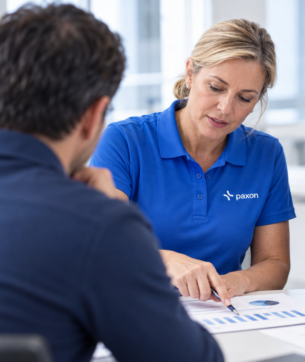 A woman in a blue Paxon polo shirt points to data charts on a document while talking to a man.
