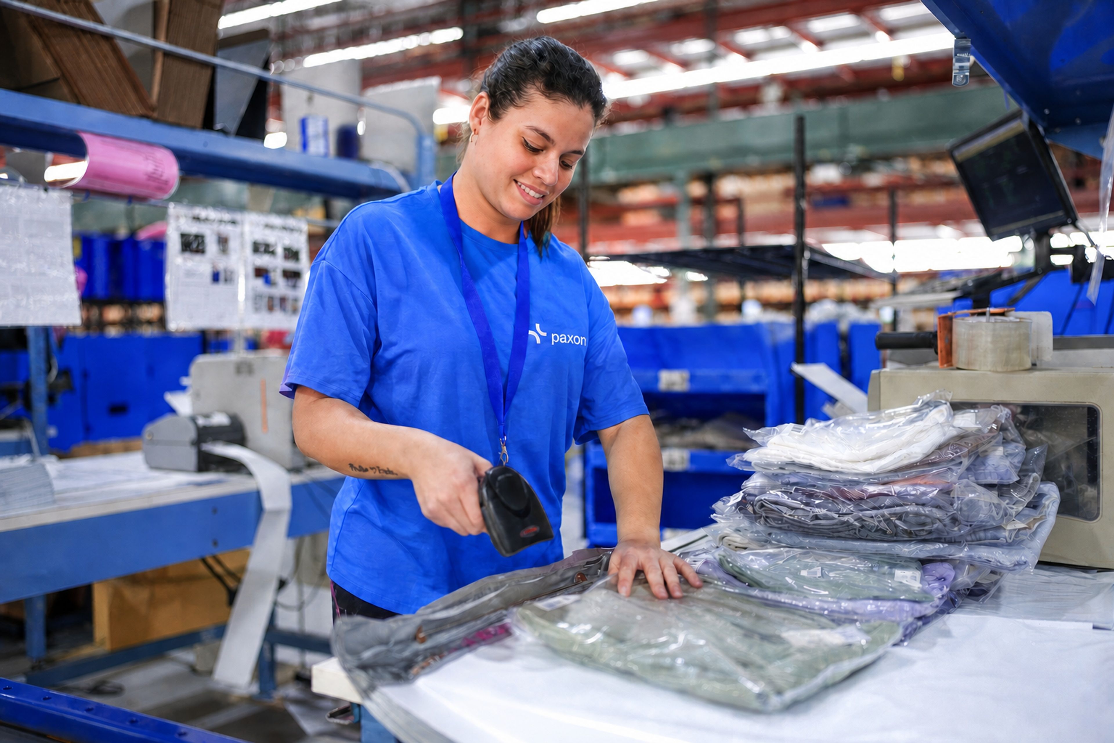Une femme en t-shirt bleu Paxon sourit en scannant un article emballé sous film plastique dans un entrepôt.