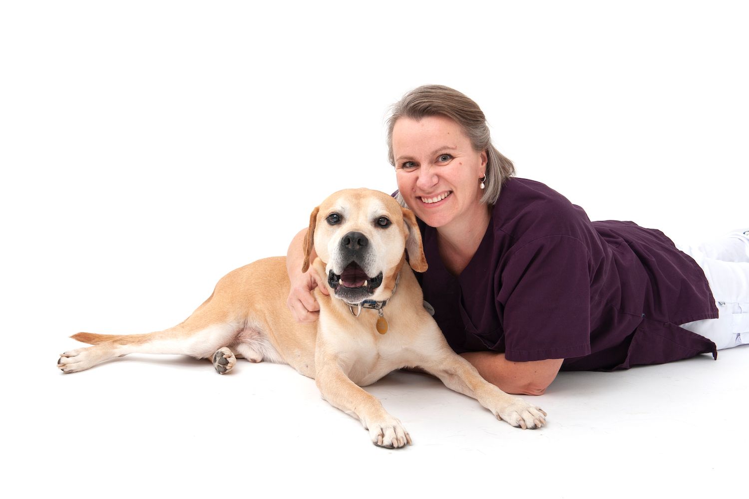 Vibeke Fjeld Harlan, veterinær ved Tønsberg Dyreklinikk, ligger på gulvet inntil en lys hund og holder rundt den, mot hvit bakgrunn