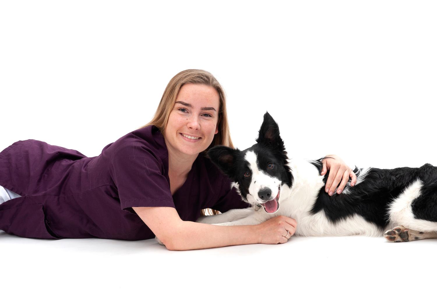 Kristine Sofie Roen, veterinær ved Tønsberg Dyreklinikk, ligger på gulvet inntil en svart og hvit hund mot hvit bakgrunn