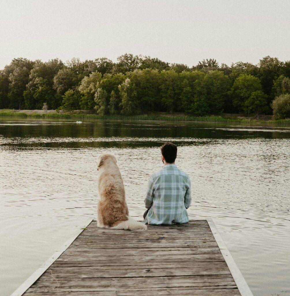 Mann og golden retriever sitter side om side ytterst på en brygge og ser ut over et stille vann