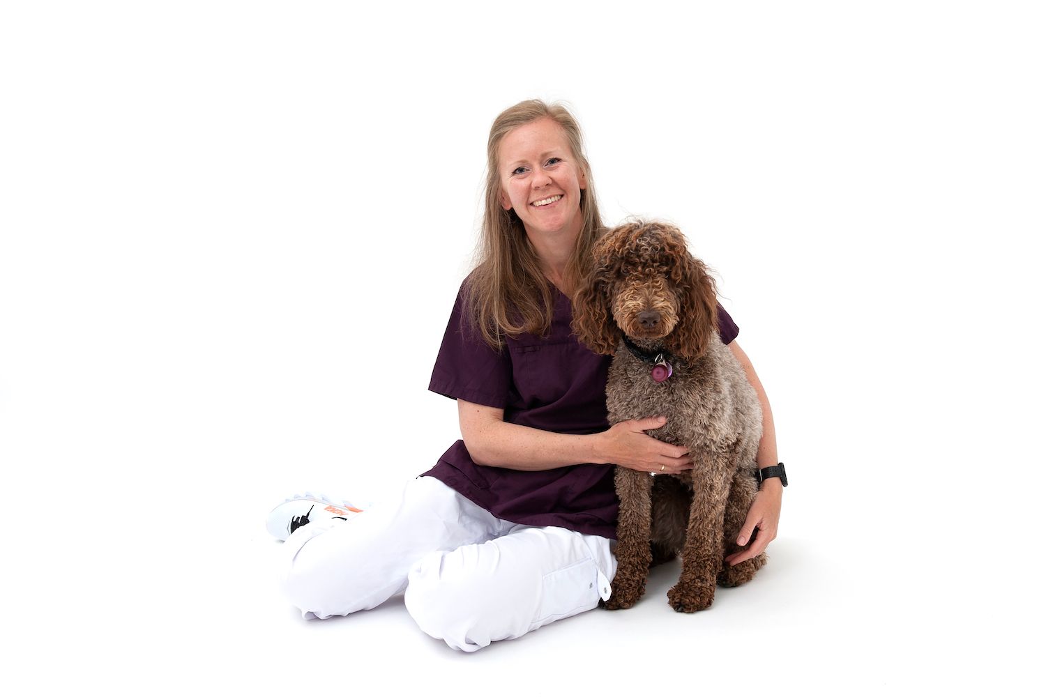 Ingvild Bjønnes Johannessen, veterinær ved Tønsberg Dyreklinikk, sitter på gulvet med armen rundt en brun hund med krøllete pels, mot hvit bakgrunn