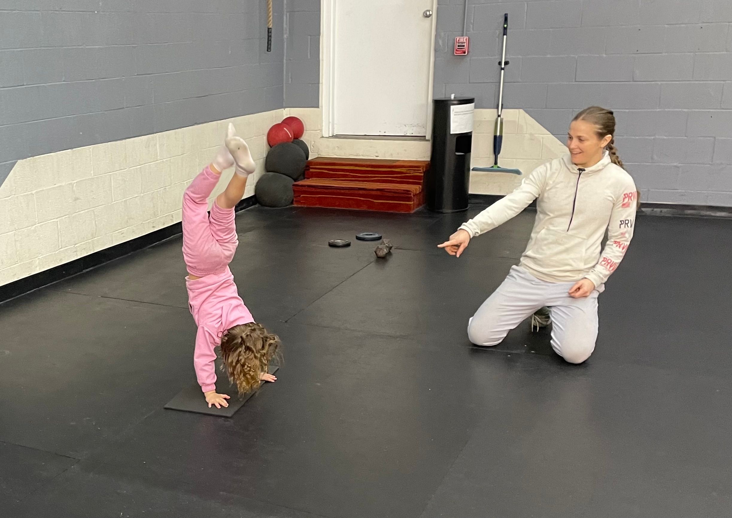 A young girl practices a handstand during a youth fitness class at Mosaic Fitness Center while a coach kneels nearby and provides guidance.