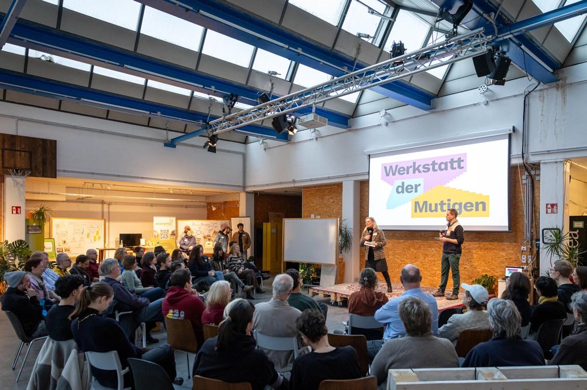 Die Eröffnungsrede bei der Werkstatt der Mutigen mit dem Publikum im Vordergrund und der Bühne und den Redner*innen im Hintergrund.