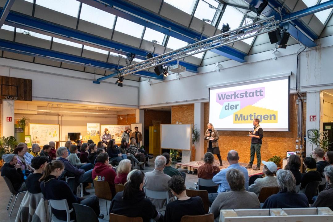Die Eröffnungsrede bei der Werkstatt der Mutigen mit dem Publikum im Vordergrund und der Bühne und den Redner*innen im Hintergrund.