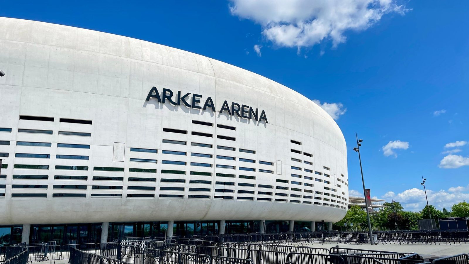 Façade blanche et courbe de l'Arkéa Arena à Floirac sous un ciel bleu, avec les barrières d'accueil du public au premier plan