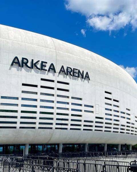 Façade blanche et courbe de l'Arkéa Arena à Floirac sous un ciel bleu, avec les barrières d'accueil du public au premier plan