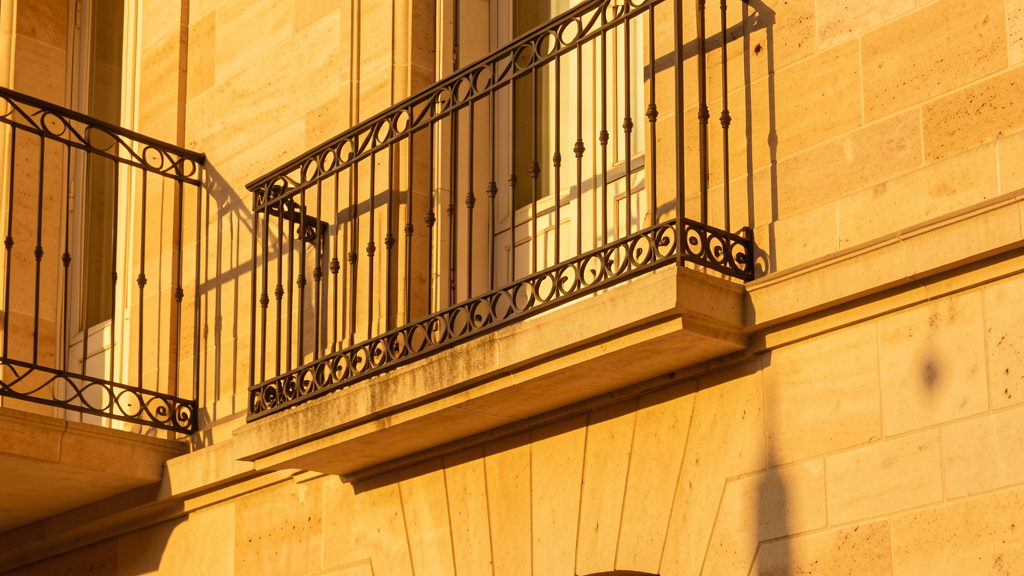 Ombres géométriques de ferronnerie bordelaise projetées sur une façade de pierre calcaire, baignées de lumière dorée