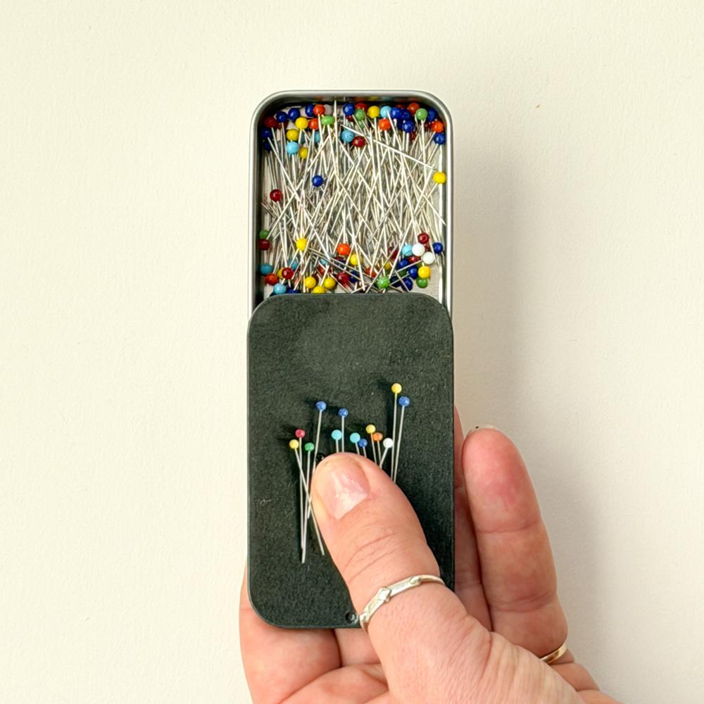 Ei hand held ei lita metallboks med fargerike knappenåler, og nokre nåler sit fast i ei svart pute framfor boksen