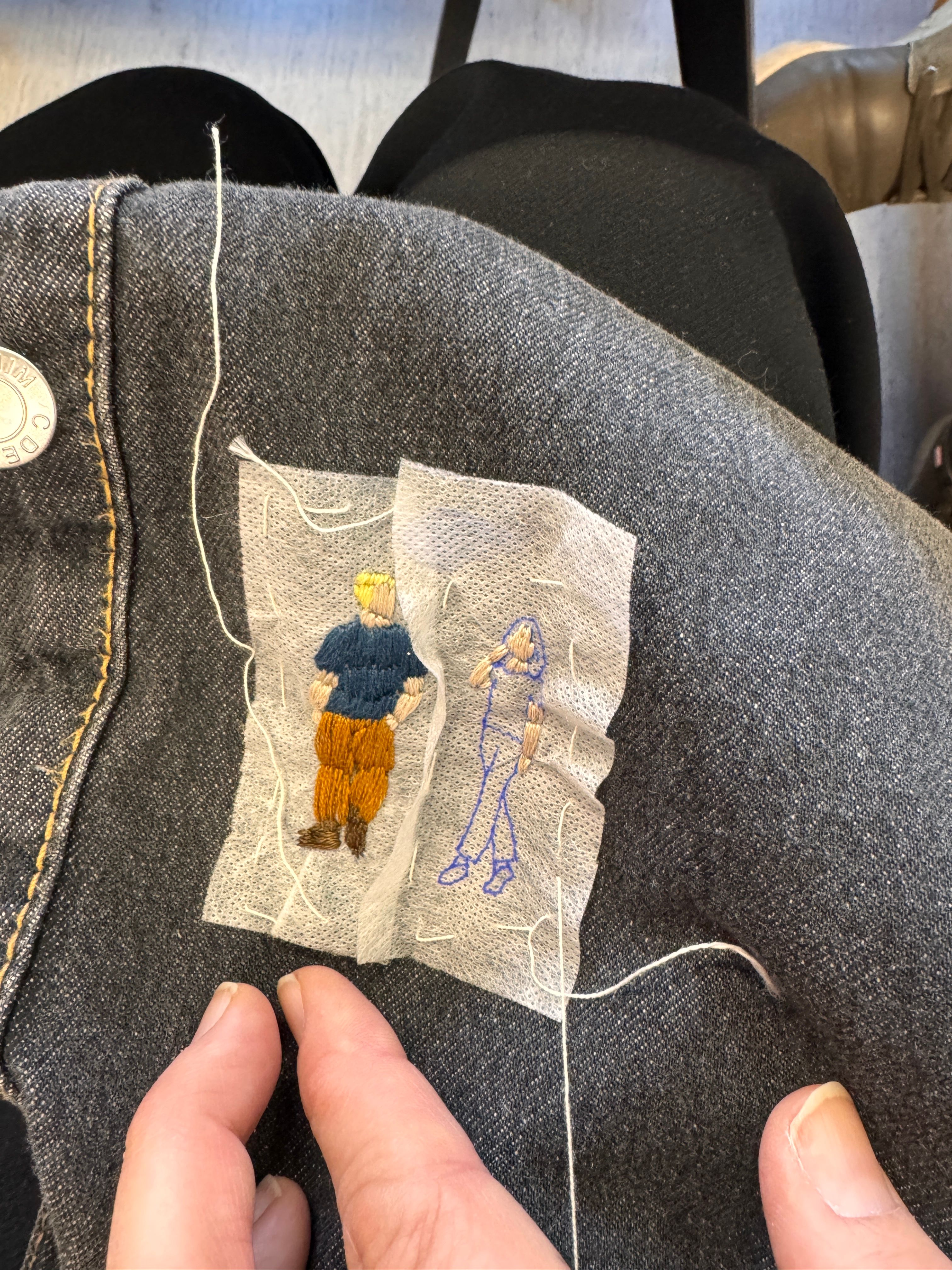 Hender som broderer ein liten person med fargerikt garn på eit mørkt klesplagg med eit tynt mellomleggstoff