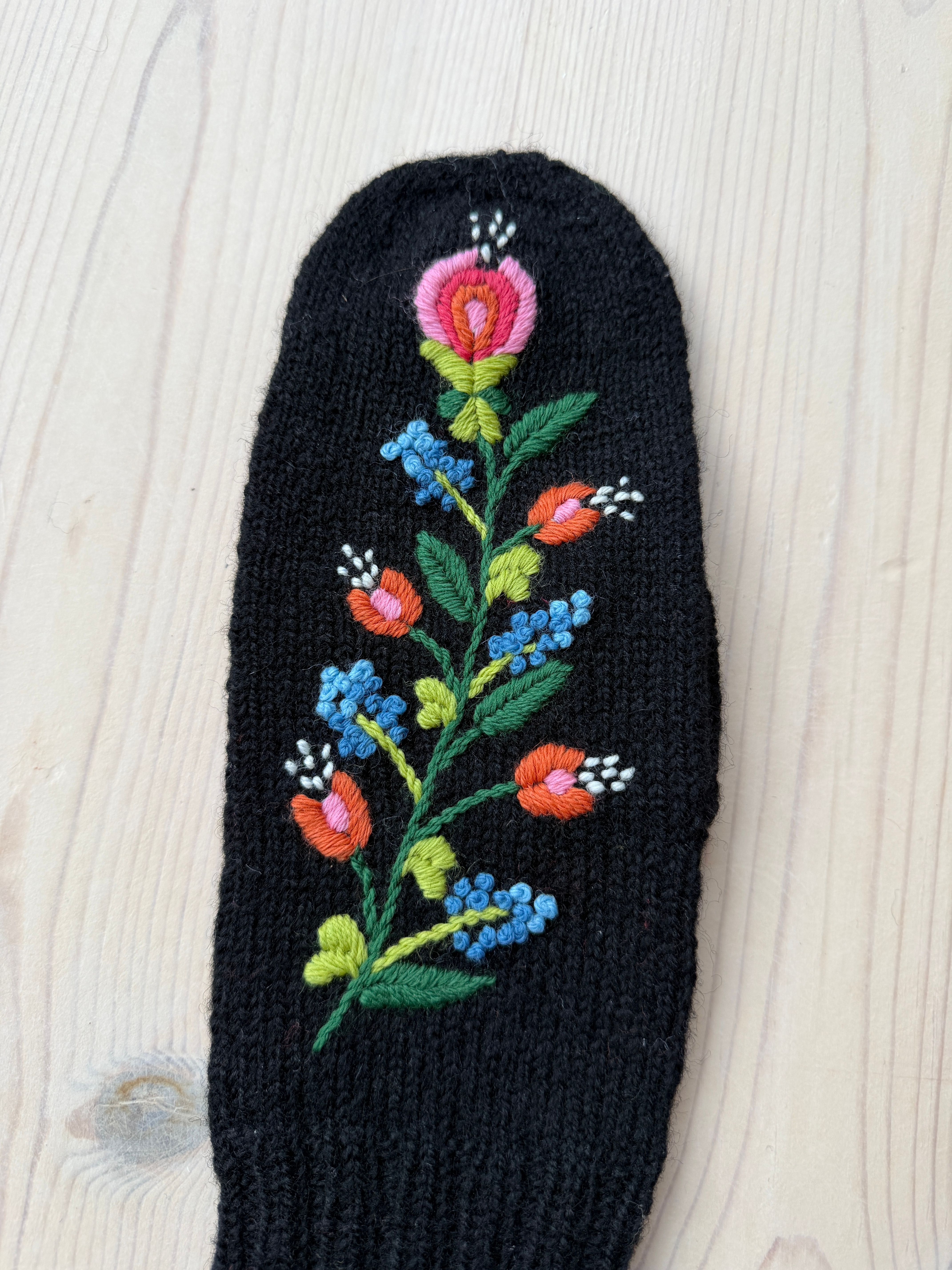 Svart strikka vott brodert med blomstrar i fargerike ullgarn på eit lyst trebord