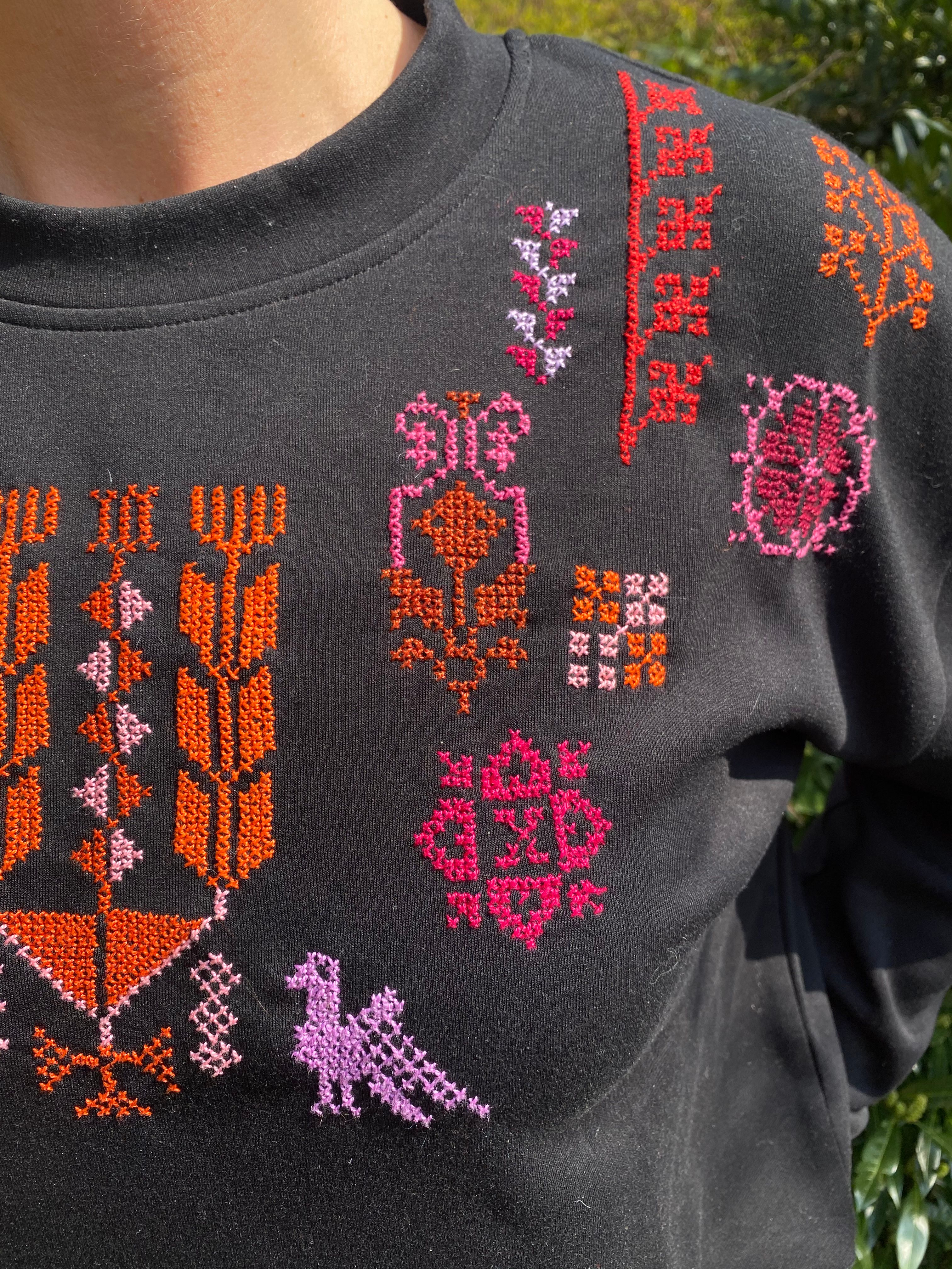 Svart genser dekorert med fargerike korsstingbroderi i raudt, oransje og rosa med tradisjonelle mønster framme på brystet