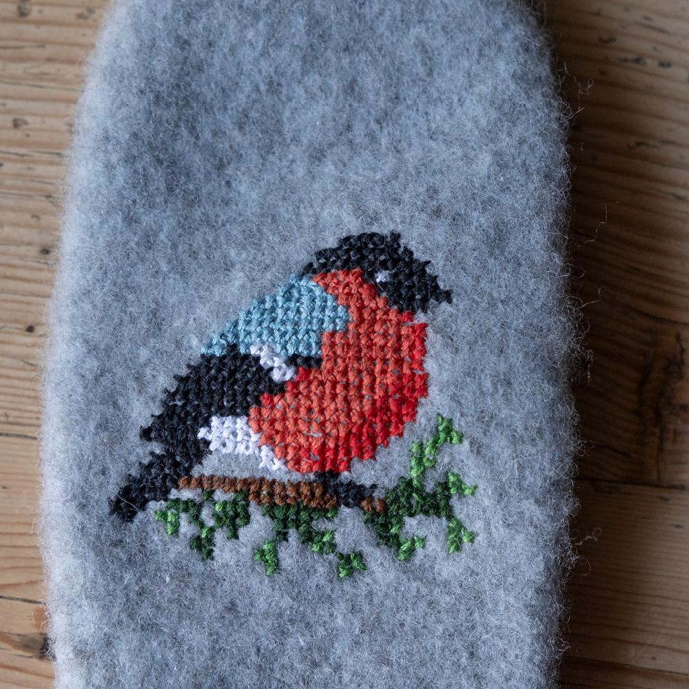 Broderi av ein dompap på ei tova grå ullflate