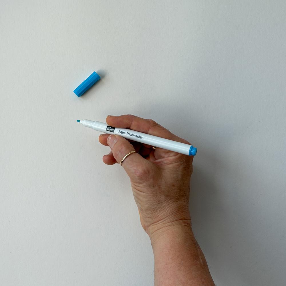 Ei hand held ein kvit og blå merkepenn med teksten Aqua-Trickmarker og hatten ligg ved sidan av mot ein kvit bakgrunn