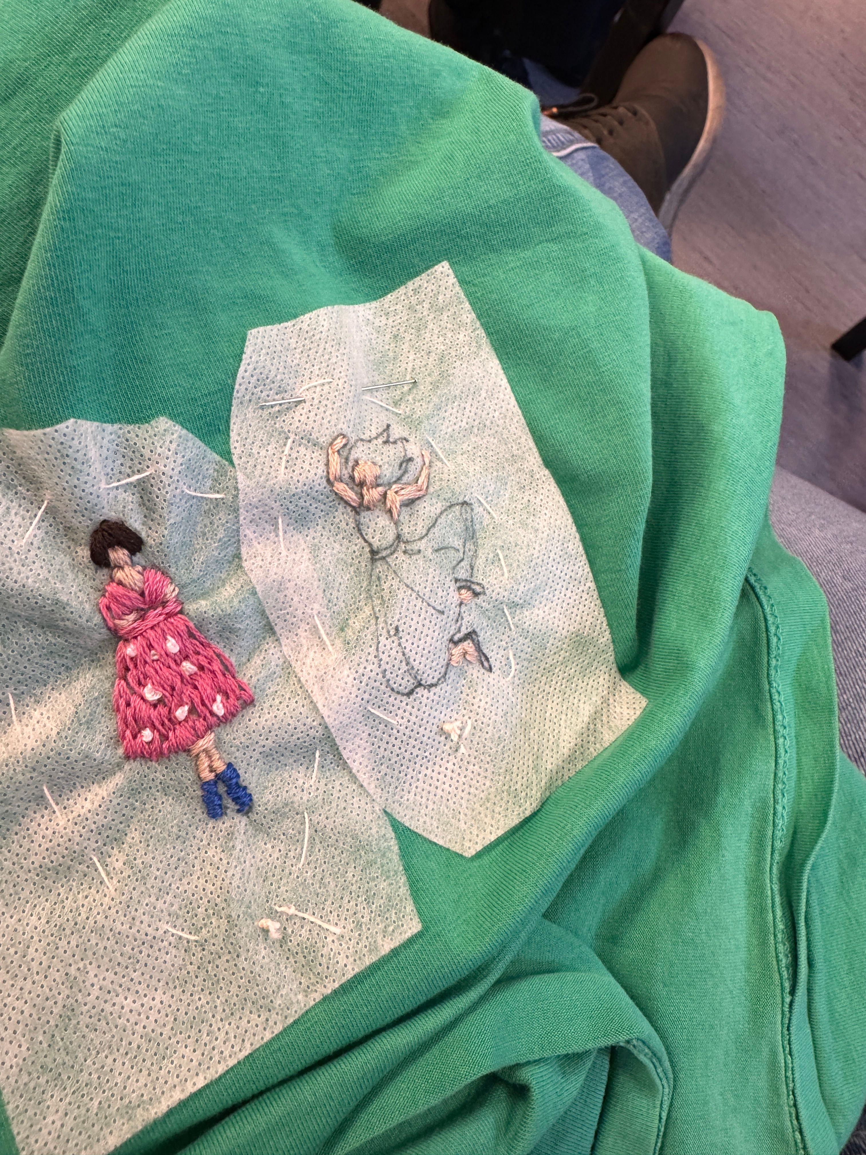 To handbroderte figurar med fargerikt garn på eit grønt plagg, festa med nålar og vattenlevande stoff