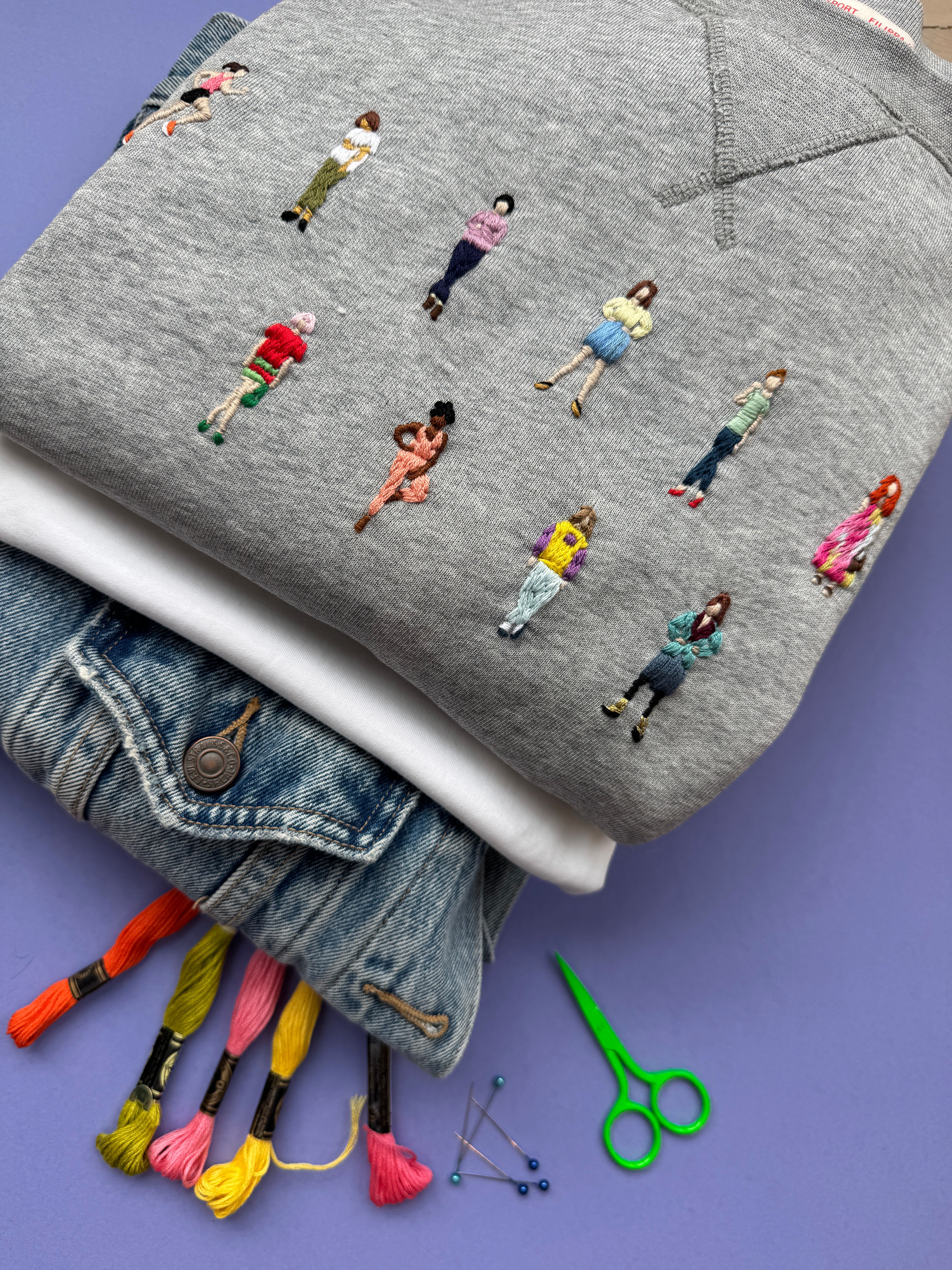 Grå genser med broderte miniatyrfolk, lagd oppå ei olajakke med broderigarn og saks attmed