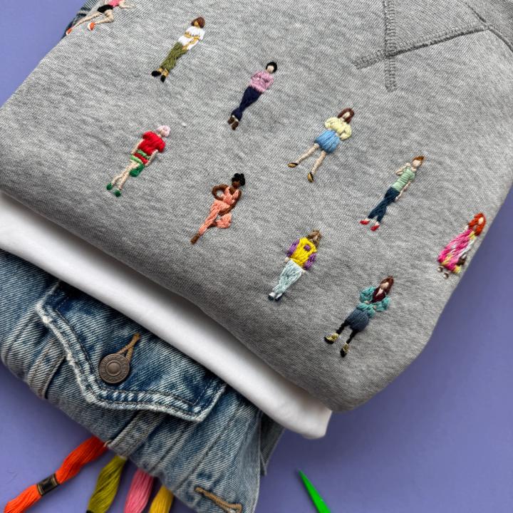 Grå genser med broderte miniatyrfolk, lagd oppå ei olajakke med broderigarn og saks attmed