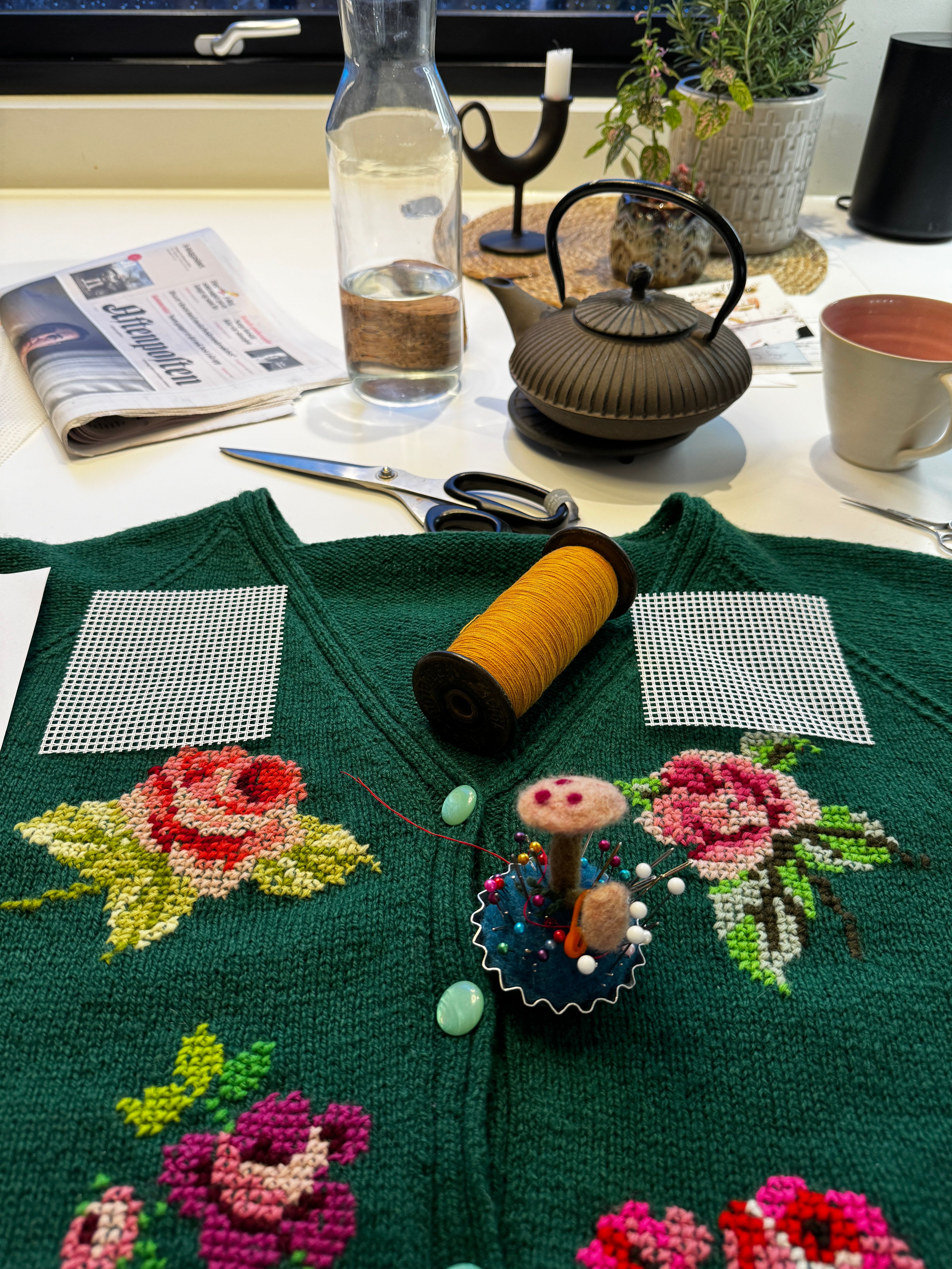 Grøn strikkejakke med broderte blomar, nålpute, gul tråd og saks på eit bord