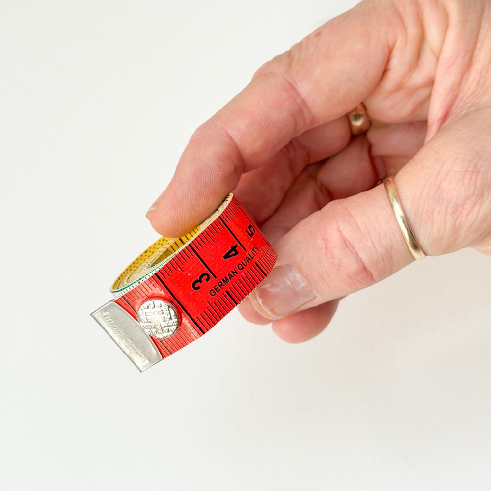 Hand som held eit lite raudt målband med trykknapp og tal frå tre til fem synleg mot ein kvit bakgrunn