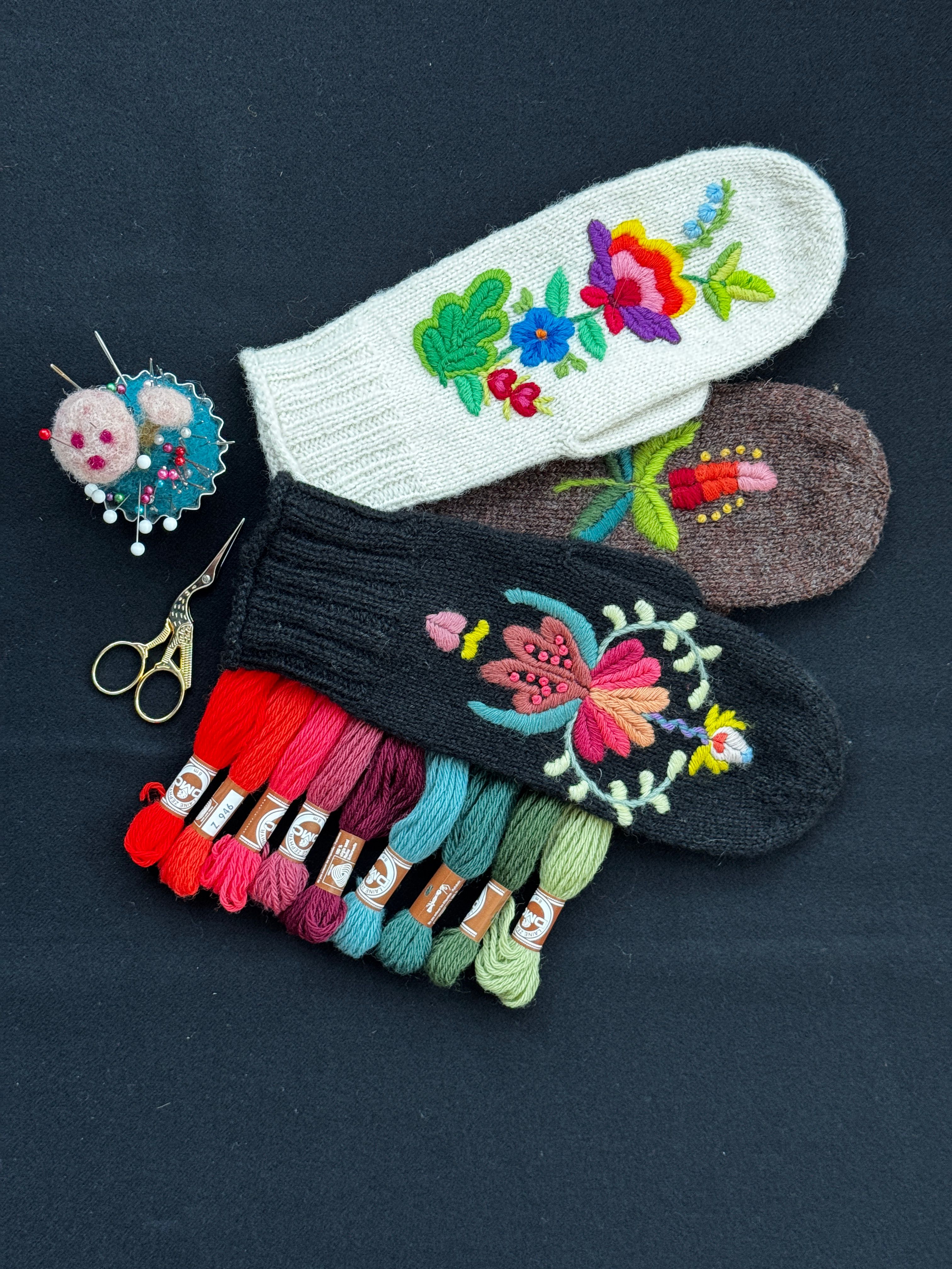 Tre broderte vottar i kvitt, brunt og svart med blomemotiv, saman med nålepute, saks og broderigarn i mange fargar