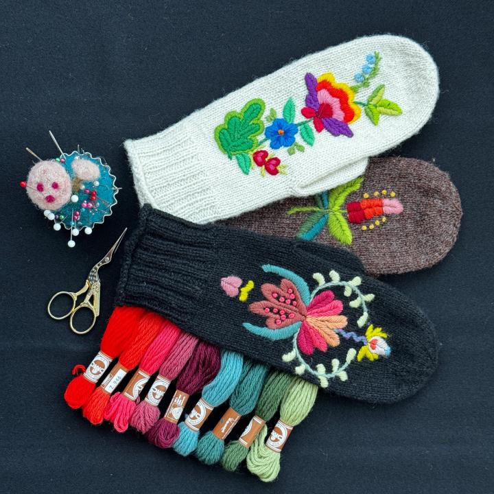 Tre broderte vottar i kvitt, brunt og svart med blomemotiv, saman med nålepute, saks og broderigarn i mange fargar