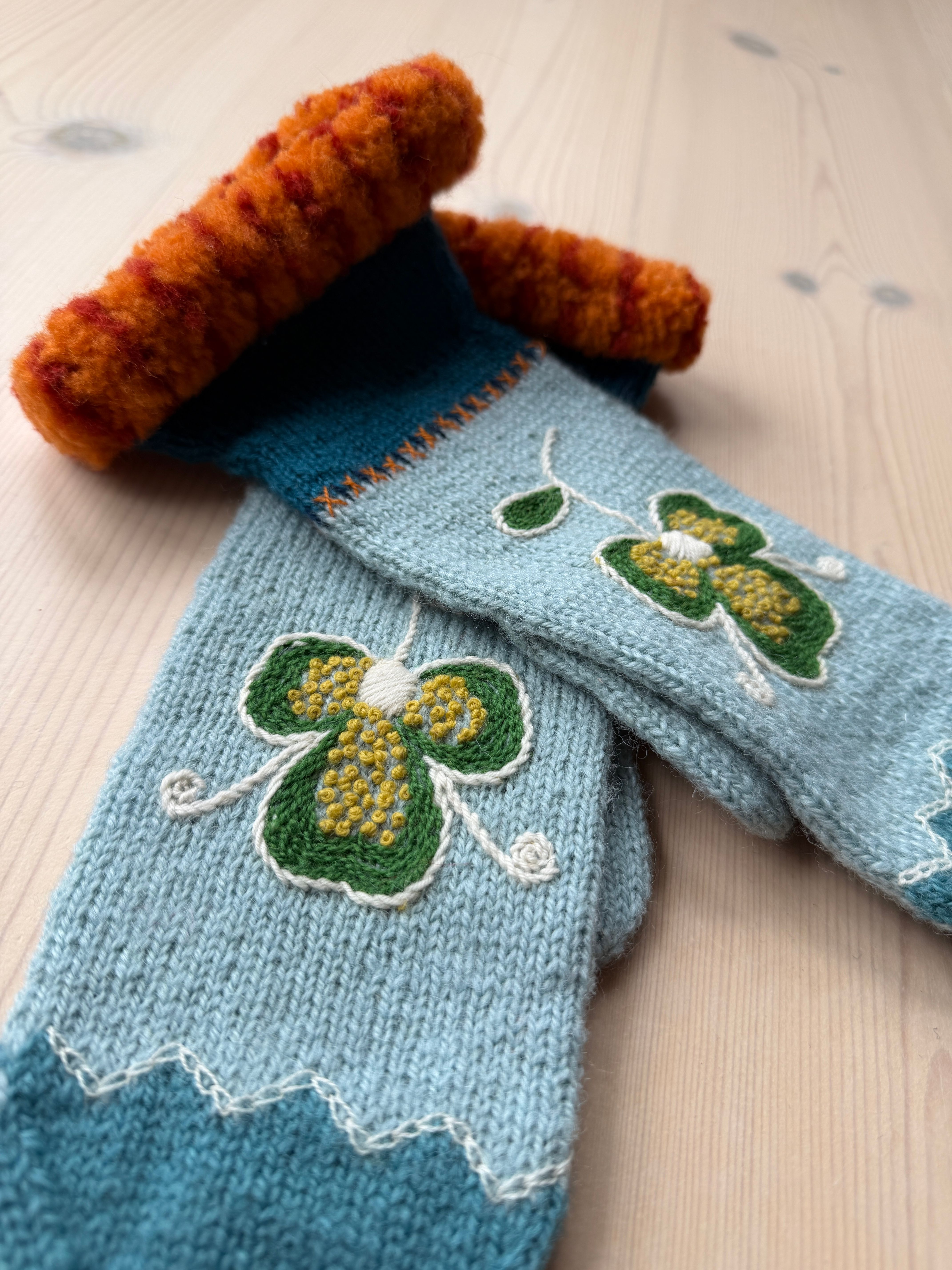 Lyseblå strikka vottar med broderte grøne blomar og gule franske knutar ligg på eit lyst trebord
