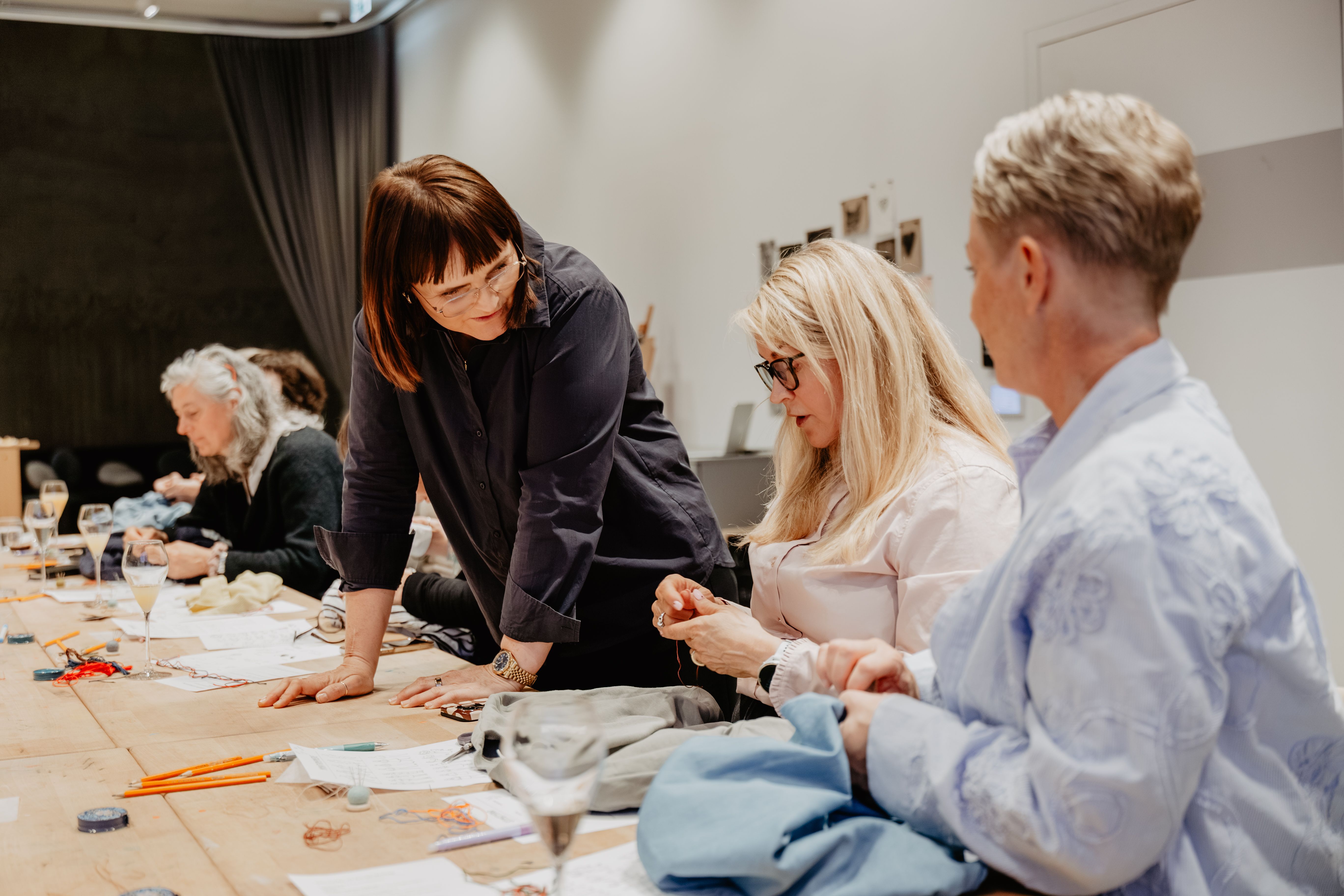 Kathrine står ved eit langbord og hjelper deltakarar med handverk og sying under eit kurs