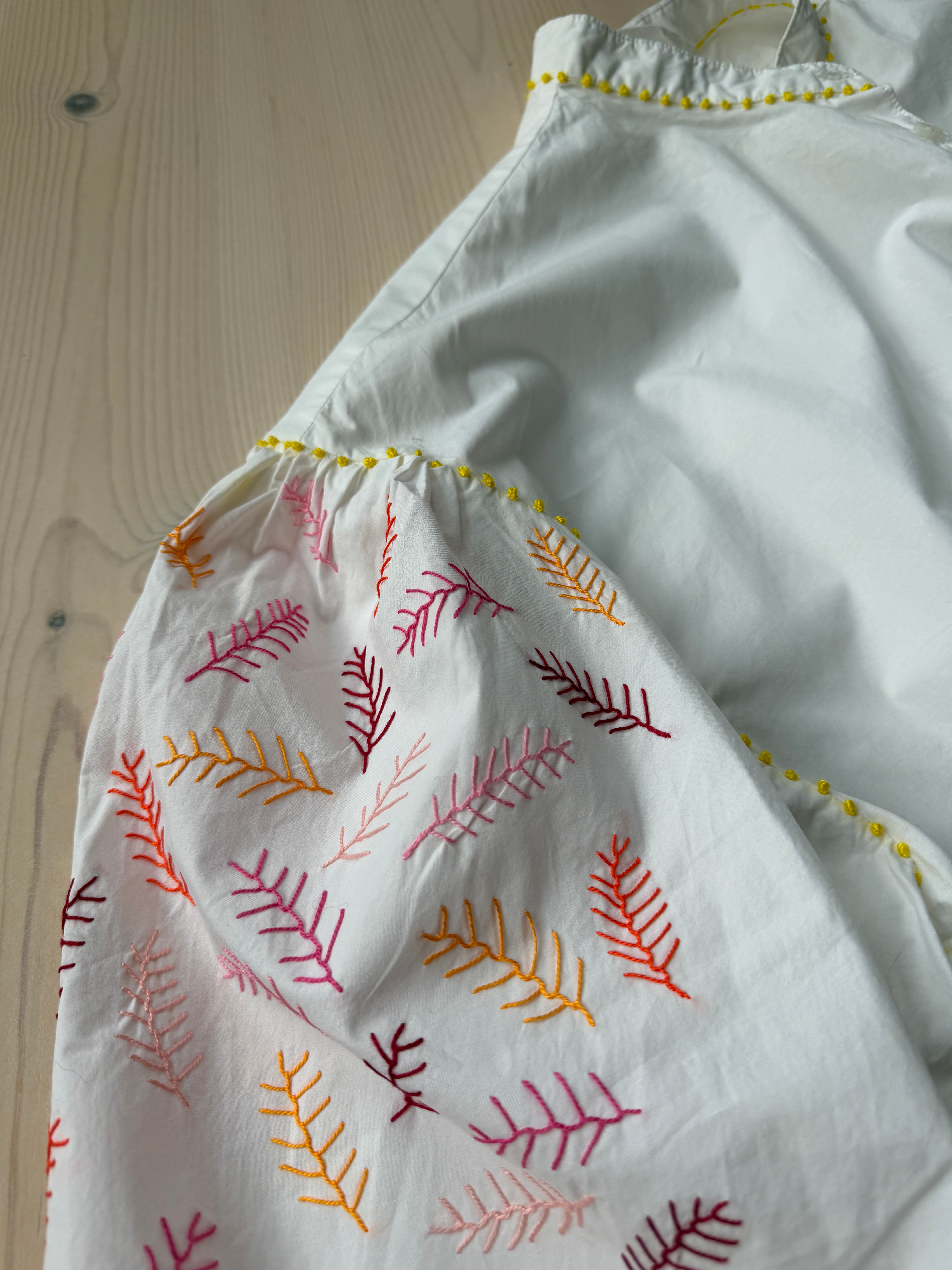 Kvit bluse med broderte lauvmønster i gult, rosa og orange og små gule franske knutar langs sømmane