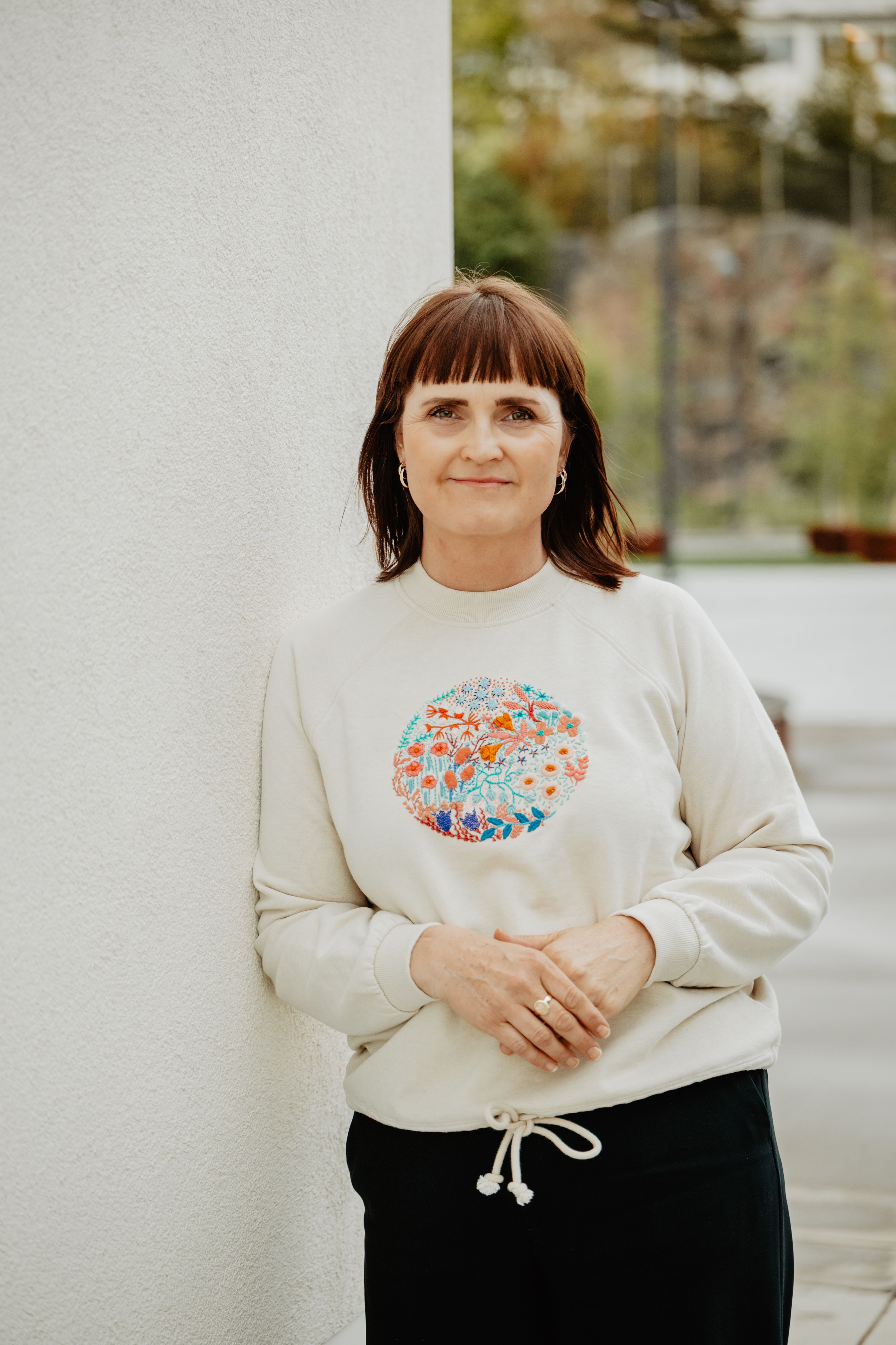 Kvinne med brunt hår står mot kvit vegg og held hendene samla framfor seg, i lys genser med fargerik handbroderi