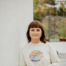 Kvinne med brunt hår står mot kvit vegg og held hendene samla framfor seg, i lys genser med fargerik handbroderi