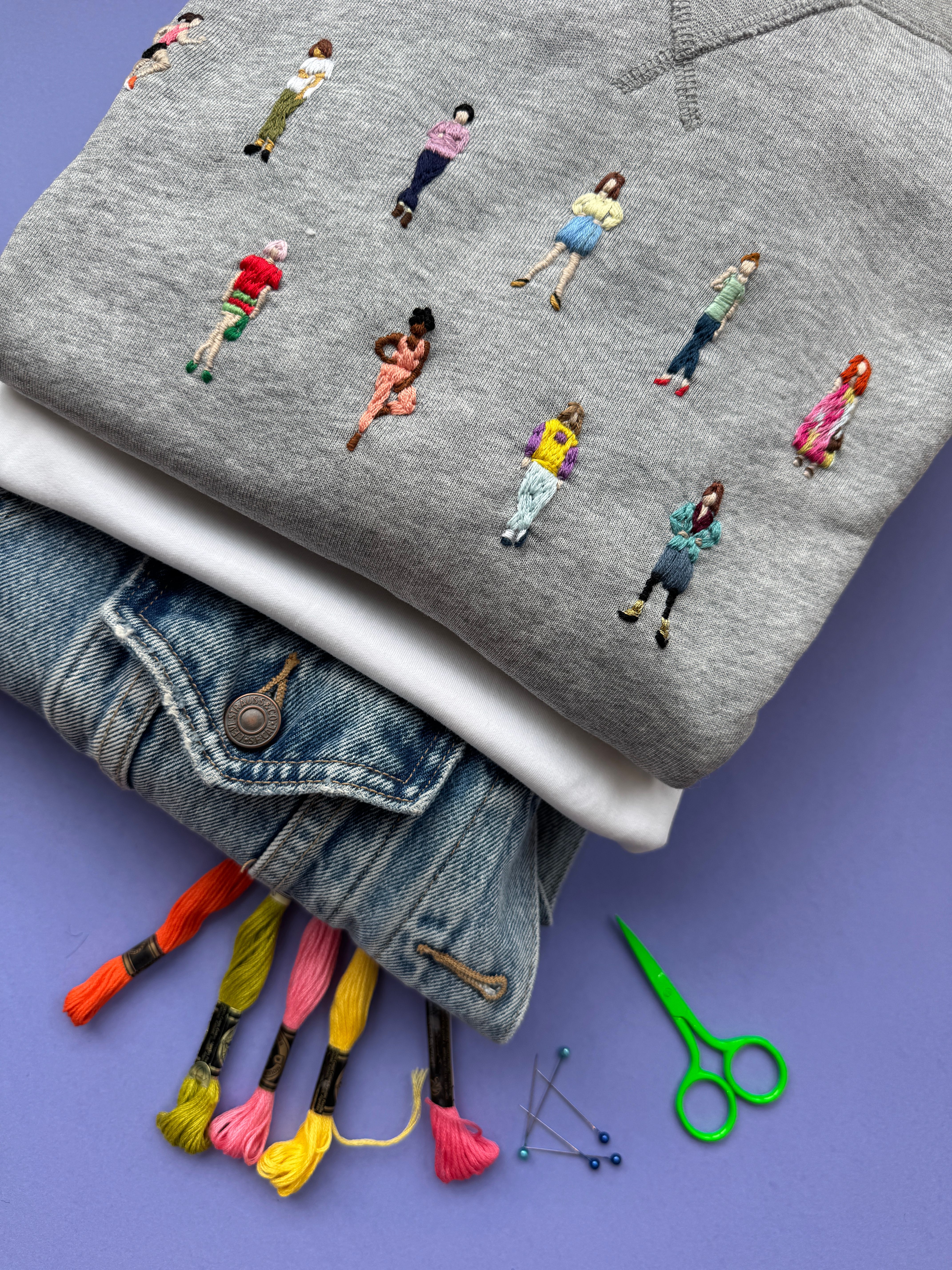 Grå genser brodert med små menneske, stabla oppå kvit skjorte og olajakke med broderigarn og saks ved sidan av