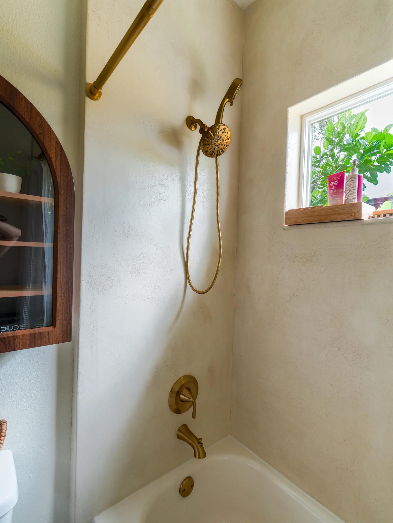 Microcement shower wall with a brass handheld shower and tub spout, window to the right