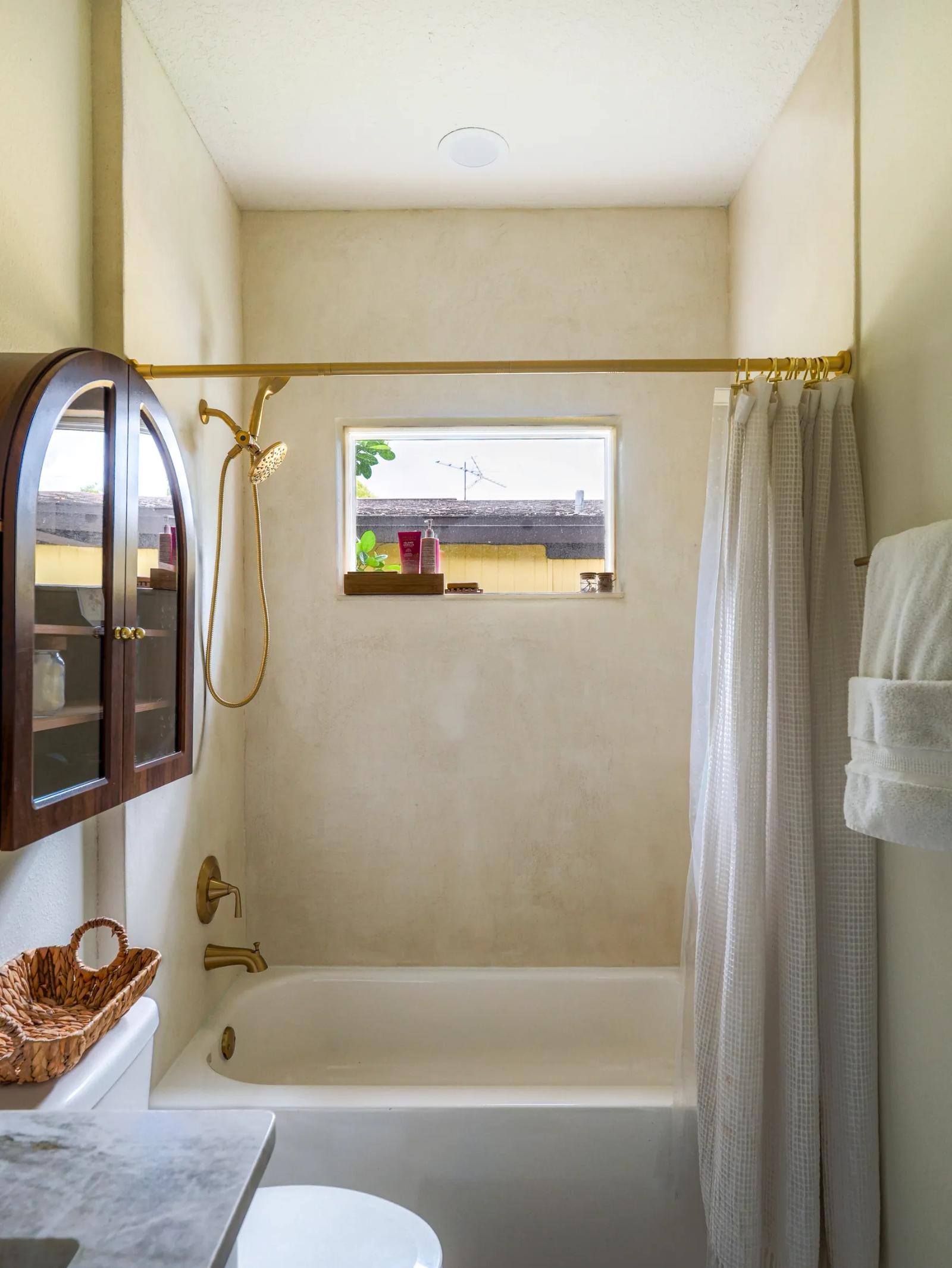 Tub alcove finished in seamless microcement, with a brass shower rod and a window above the tub