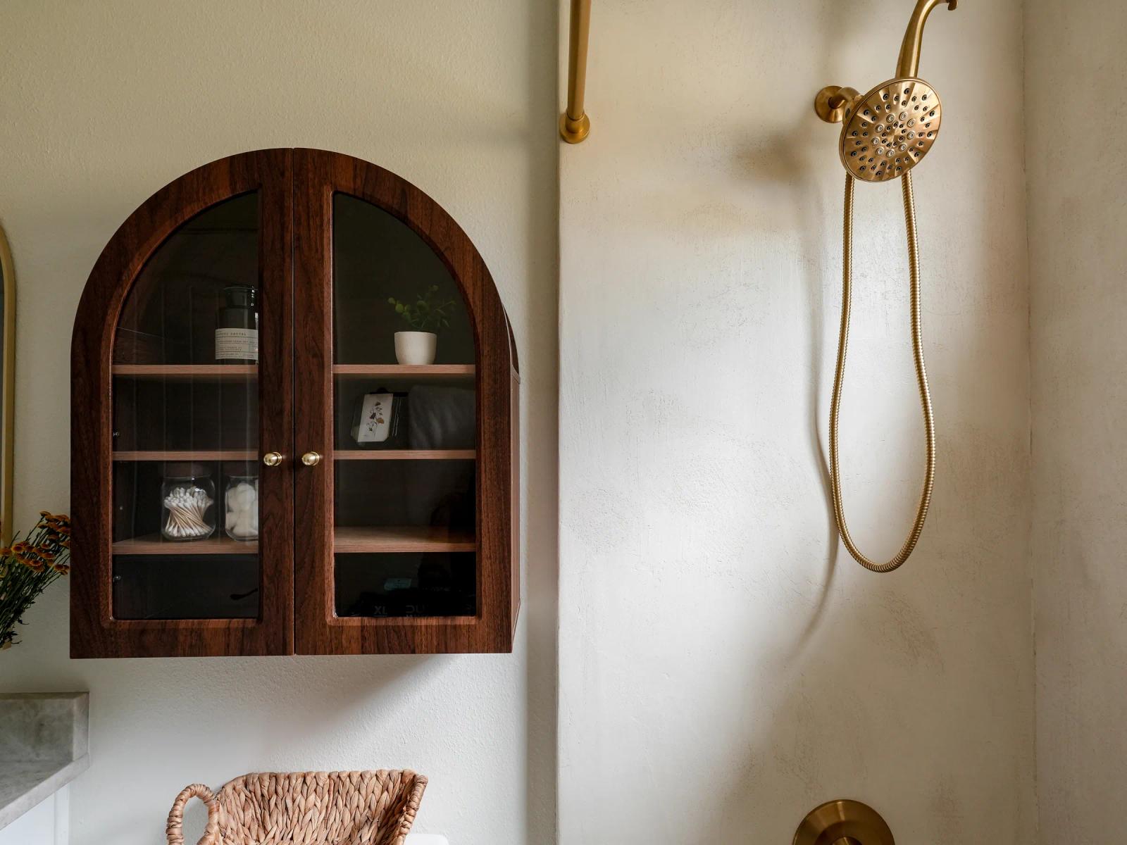 Arched dark-wood glass cabinet beside the microcement shower wall and brass shower head