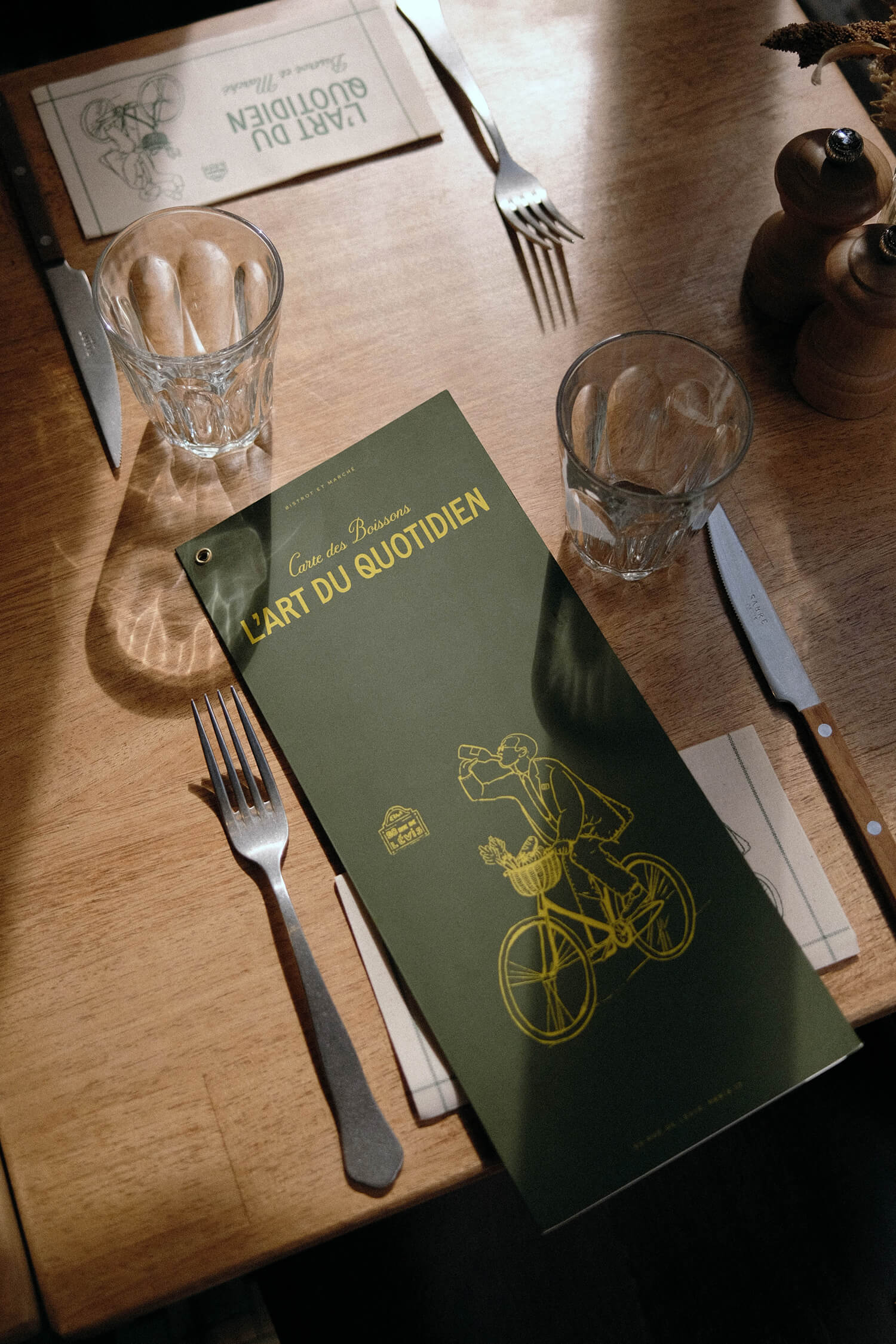 Table setting with a folded green-and-yellow menu reading "L'Art du Quotidien" beside cutlery and water glasses on a wooden table.