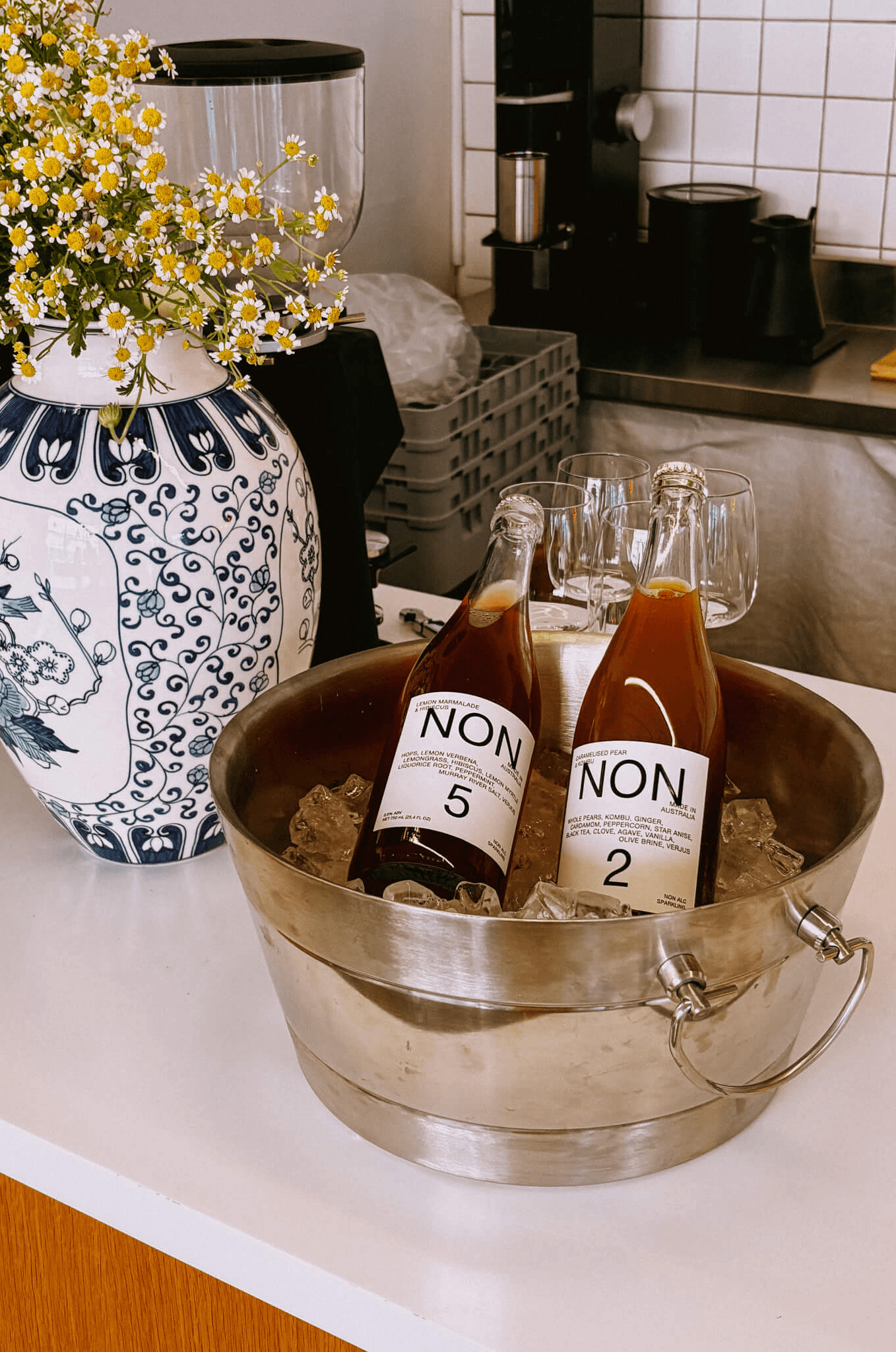 A blue-and-white vase of daisies next to an ice bucket holding two bottles of "NON" non-alcoholic drinks.