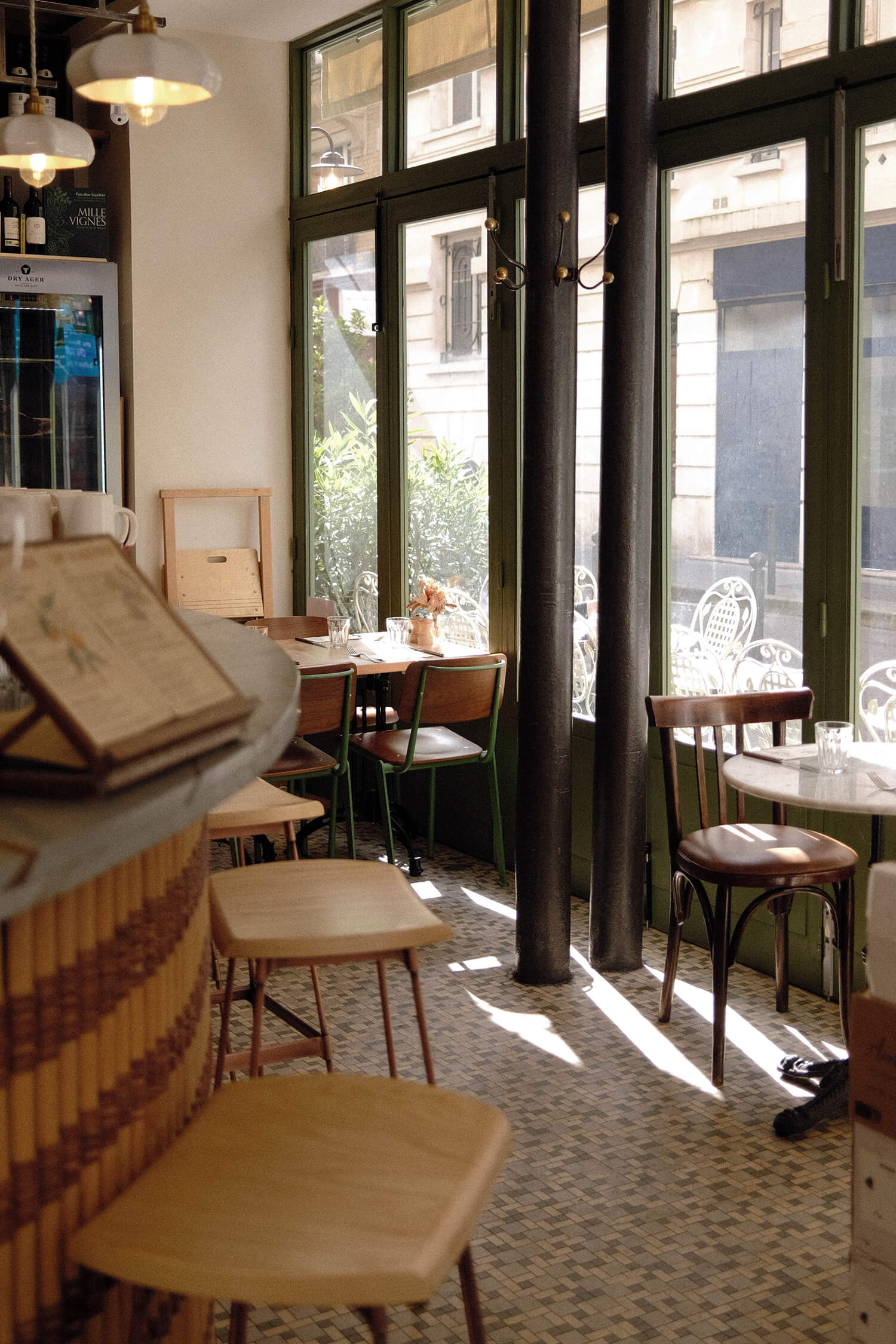 Interior view of the bistro showing wood and rattan bar stools, a mosaic tile floor, dark iron columns, and sunlit café tables by the front windows.