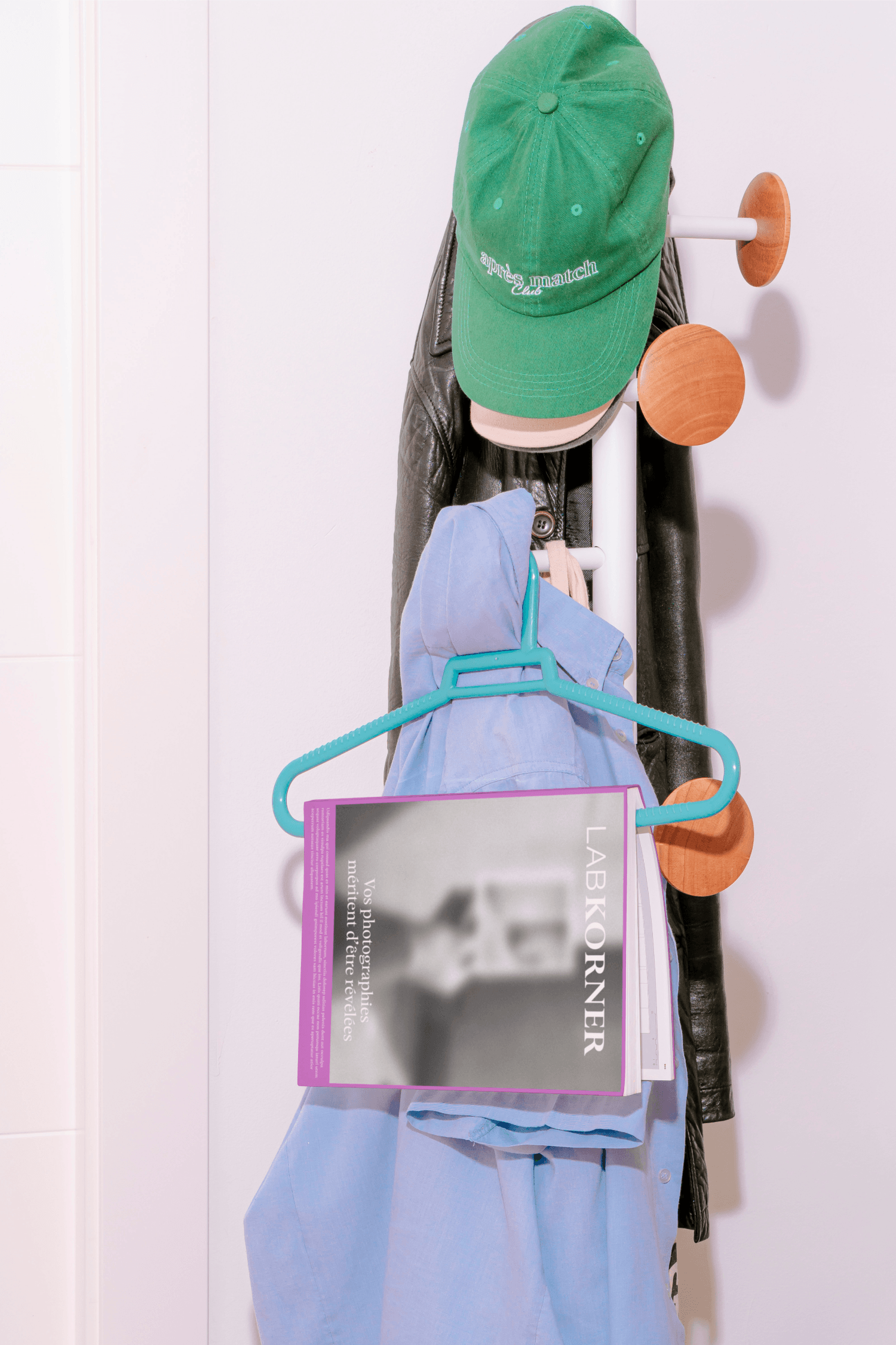 A green cap and blue jacket hanging on wall hooks beside a blue hanger, with a LabKorner magazine or brochure tucked in among the clothing.