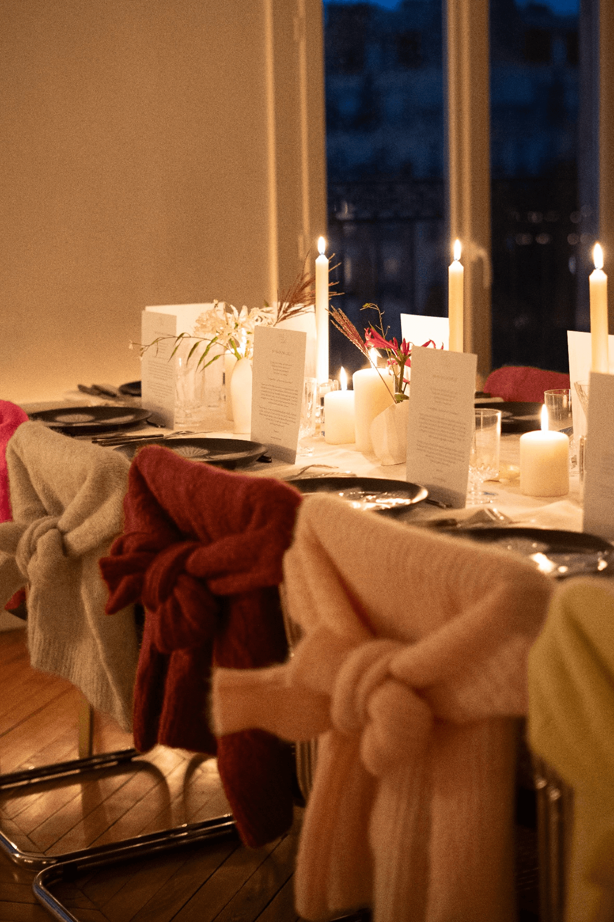 Long dinner table set with candles, place cards, and folded knit sweaters draped over each chair back, lit warmly at dusk.