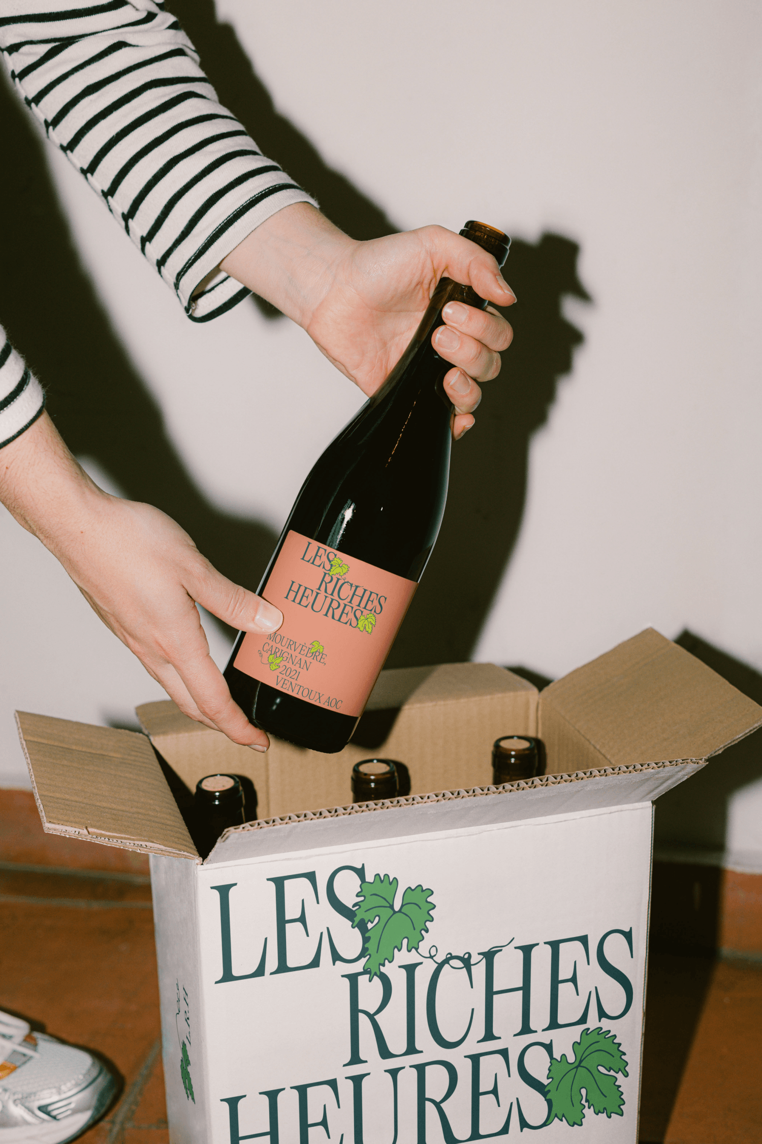 Close-up of hands placing a wine bottle into an open shipping box branded "Les Riches Heures."