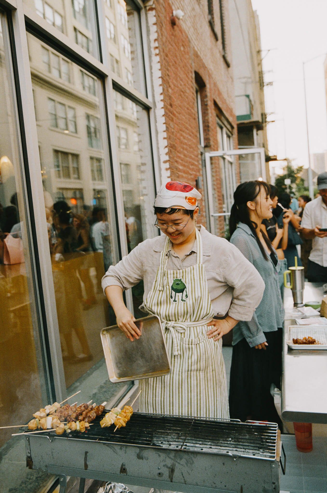 "Day with Mei" chef in an apron grilling skewers on an outdoor grill at the event.